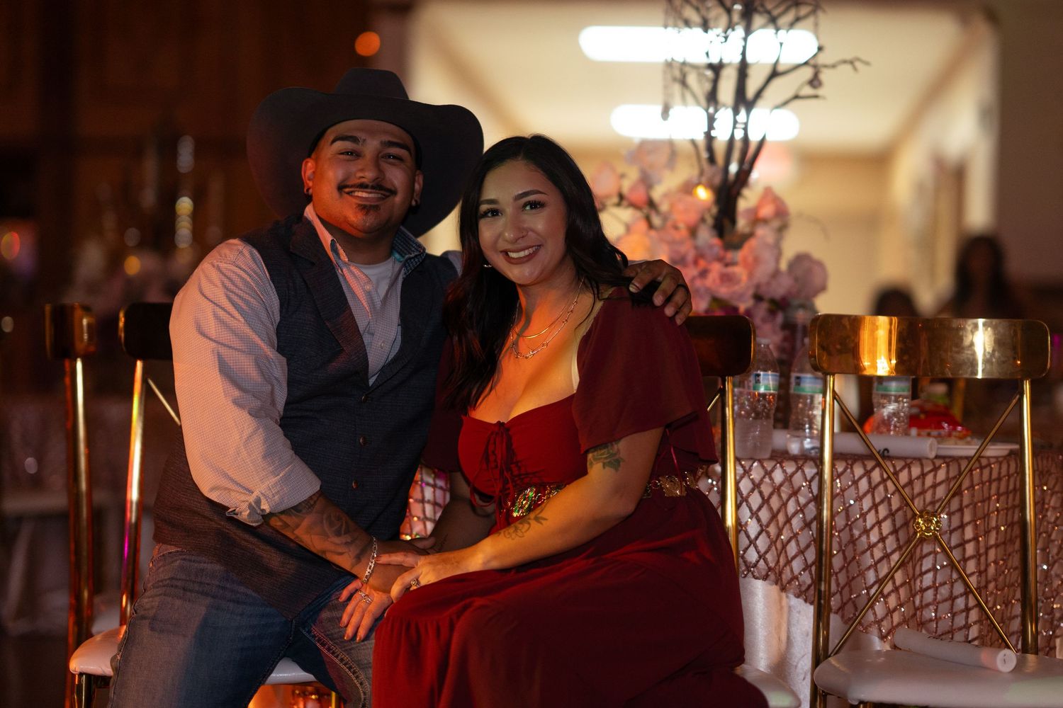 A couple in formal attire, man in cowboy hat and woman in red dress, pose together at an elegant event venue.