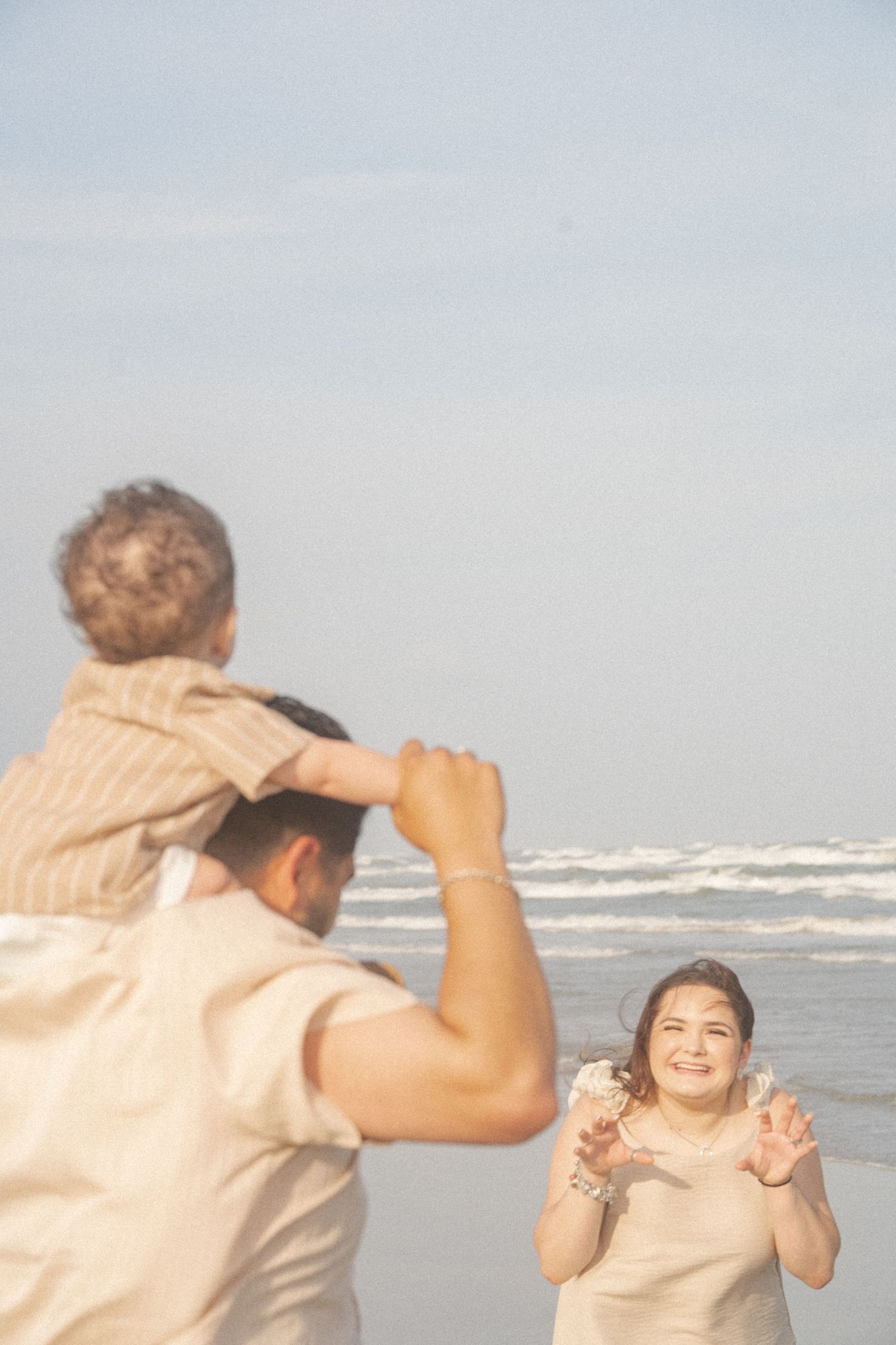 A joyful mother smiles as father lifts child on shoulders during a family beach outing.