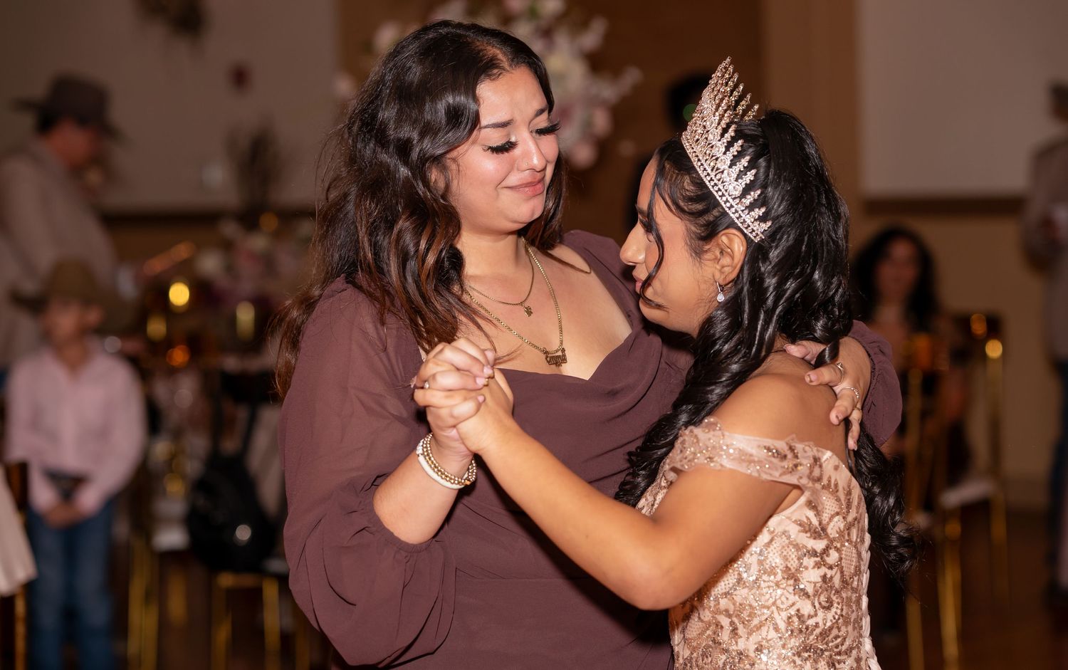 Quinceañera in elegant gold dress and tiara shares a special dance with a woman in brown at her party.