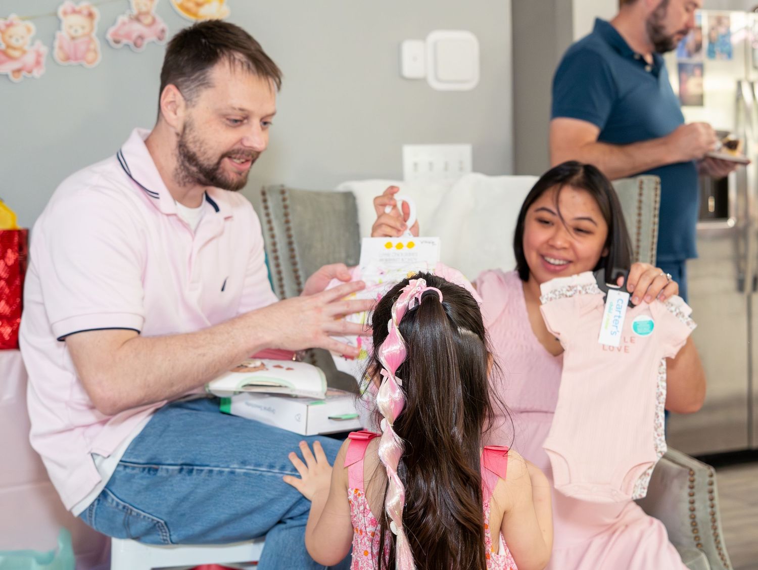 Family at a baby shower opening pink gifts, parents smiling with young daughter in a decorated room.