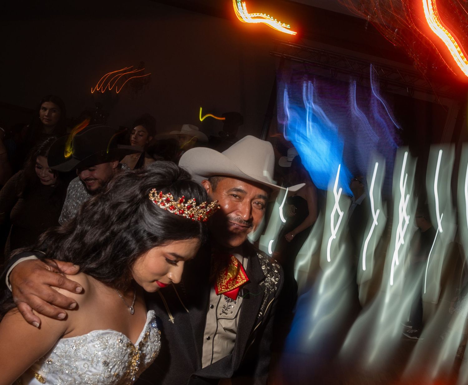 Wedding guests dance and celebrate on a dimly lit dance floor with colorful lighting effects in the background.