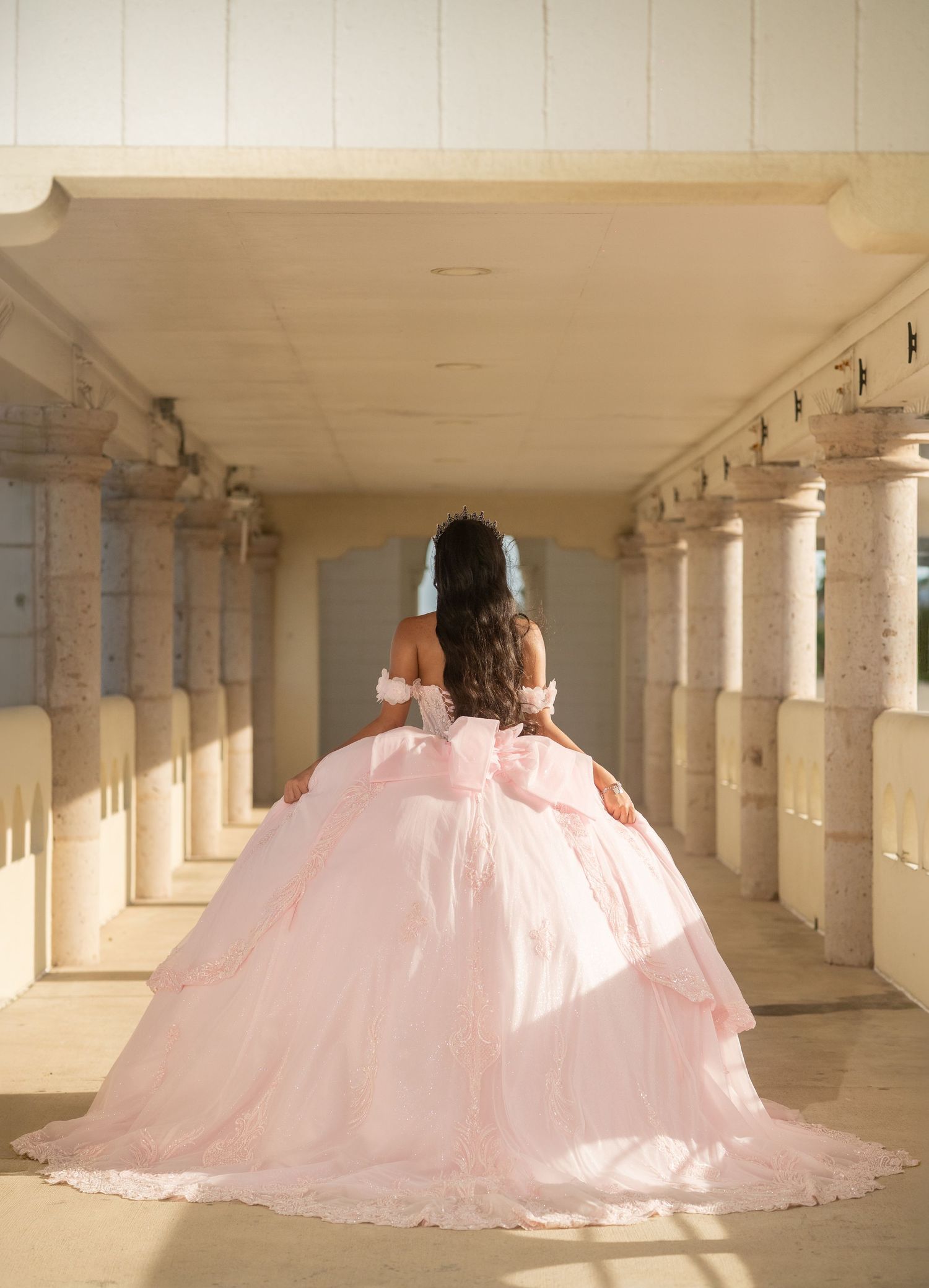 Back view of quinceañera in voluminous pink ball gown walking through sunlit columned corridor.