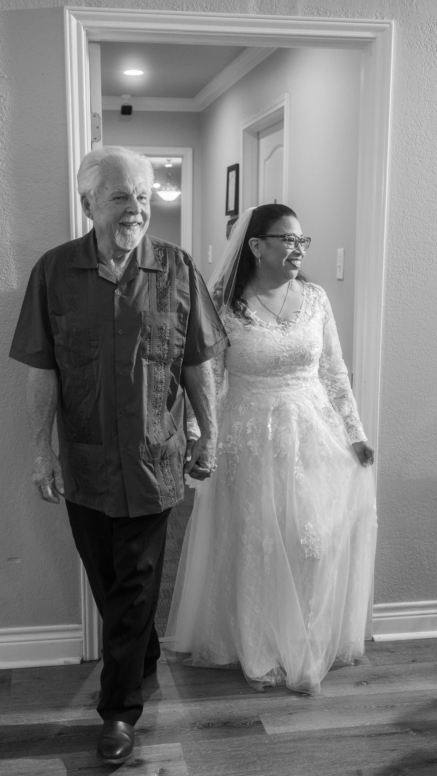 Black and white photo of a bride in lace gown holding hands with an older man walking down a hallway.