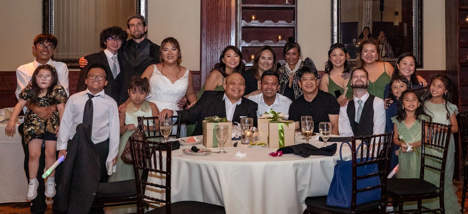 Large family group photo at a wedding reception, posing together around a decorated banquet table in an elegant venue.