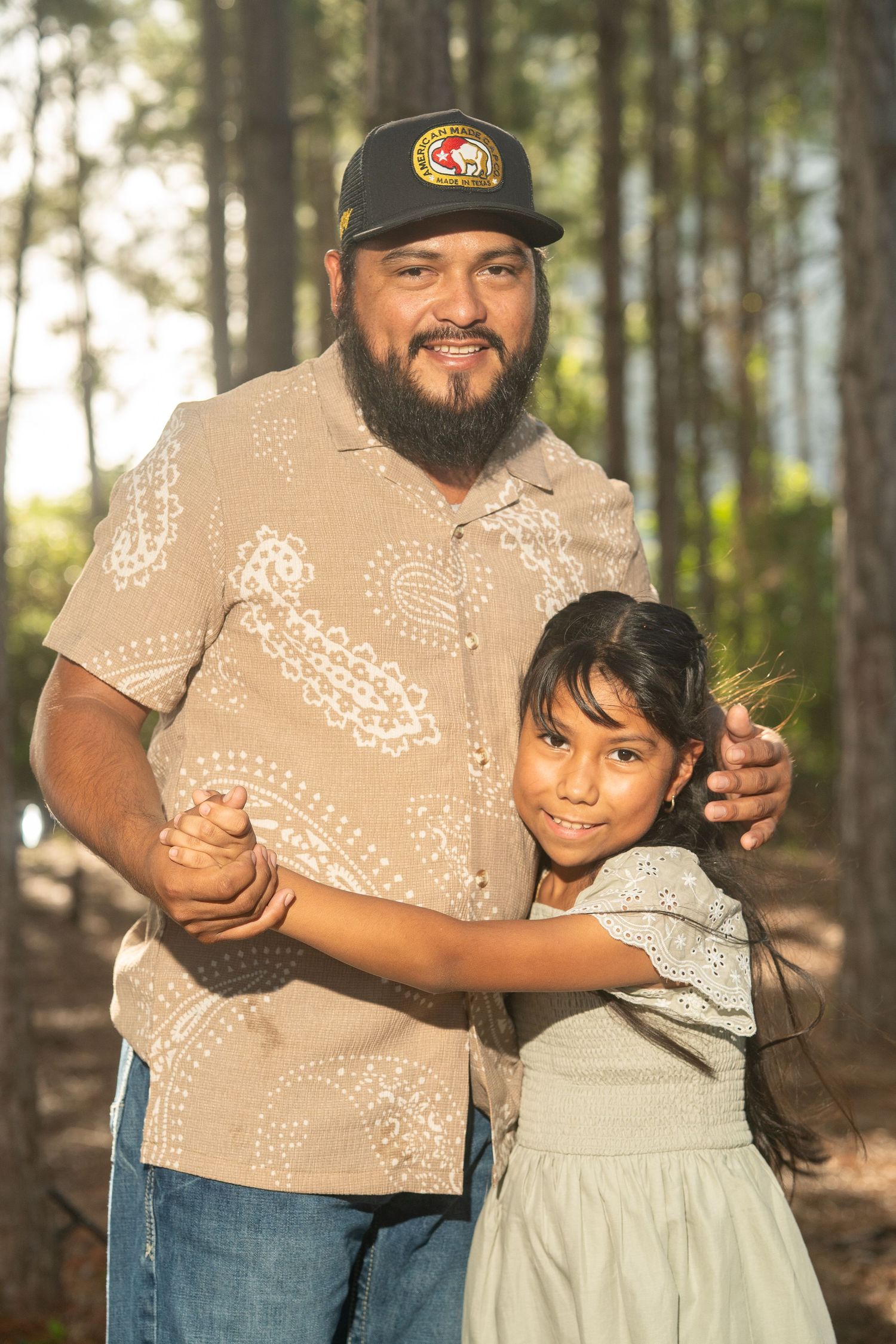 A bearded man in a cap and patterned shirt hugs a young girl in a forest setting.