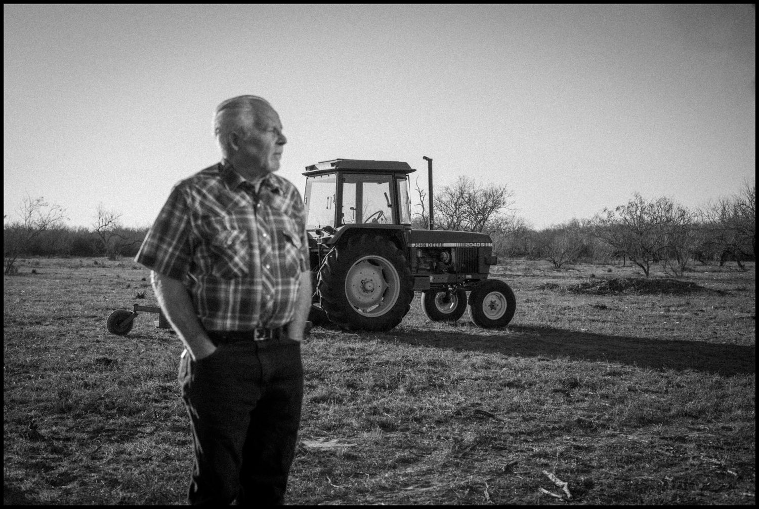 Elderly farmer stands in field beside vintage John Deere tractor, black and white rural landscape photography.