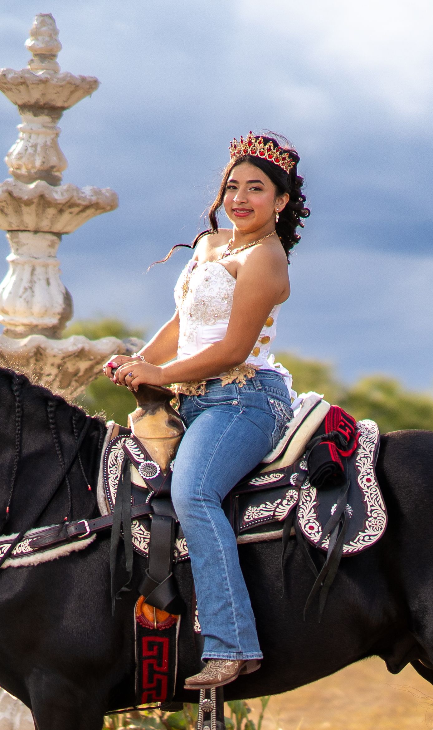 A person in jeans and white top sits on a decorated black horse saddle against a cloudy sky background.