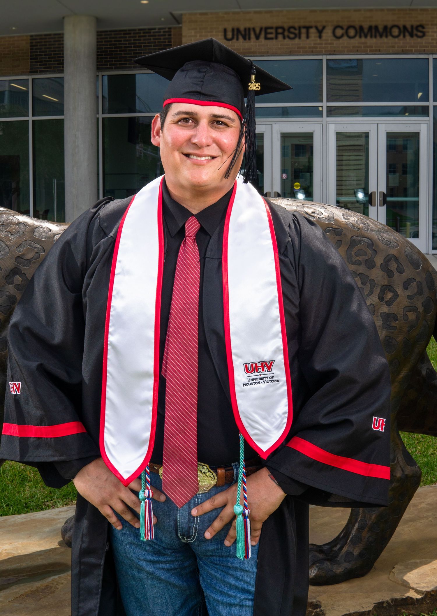 Graduate in cap and gown poses outside University Commons building wearing red tie and ceremonial stole.