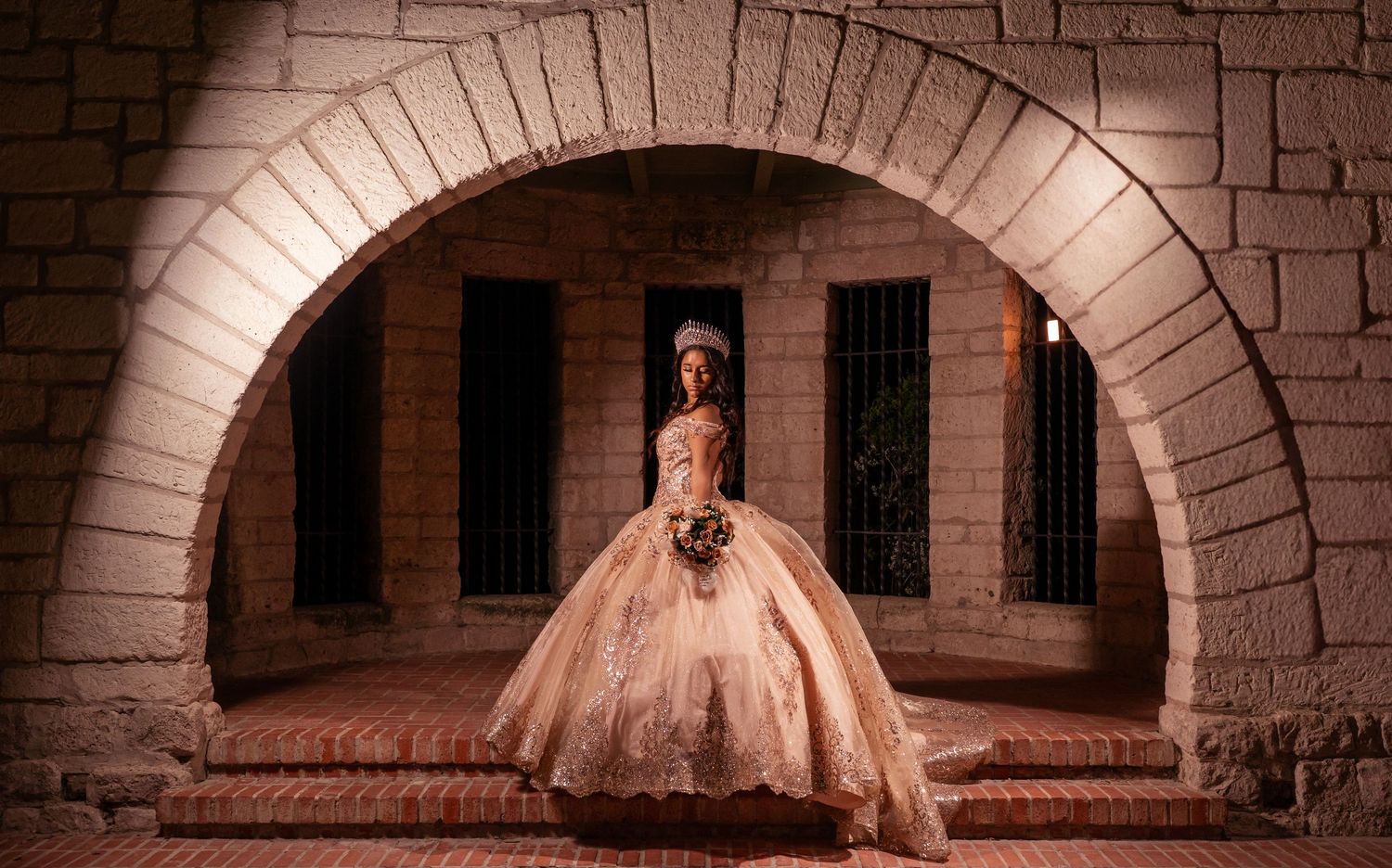 A figure in an elegant champagne-colored ballgown sits gracefully under a stone archway in a historic corridor.