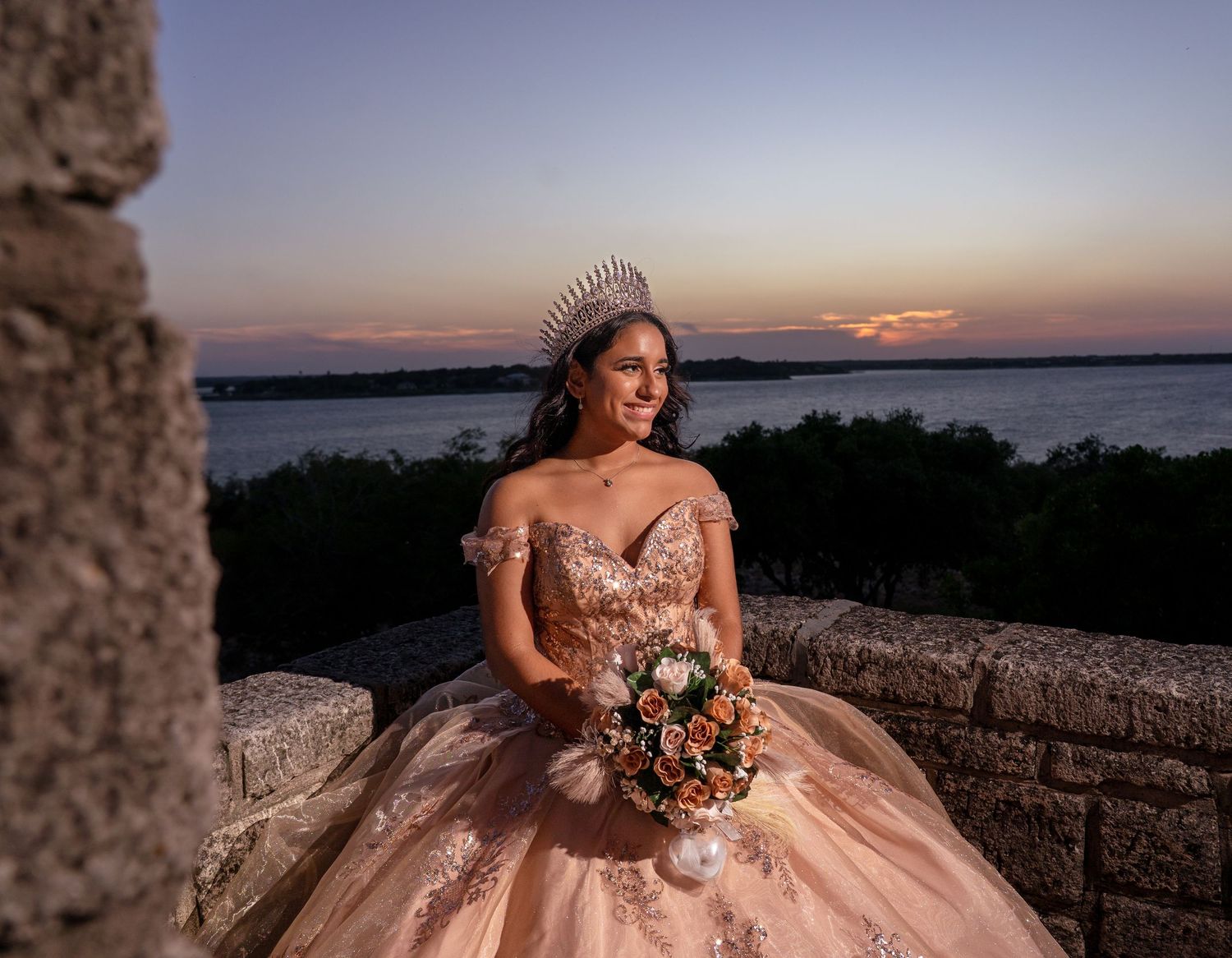A person in a blush pink ball gown poses at sunset against a stone wall overlooking a scenic waterfront view.
