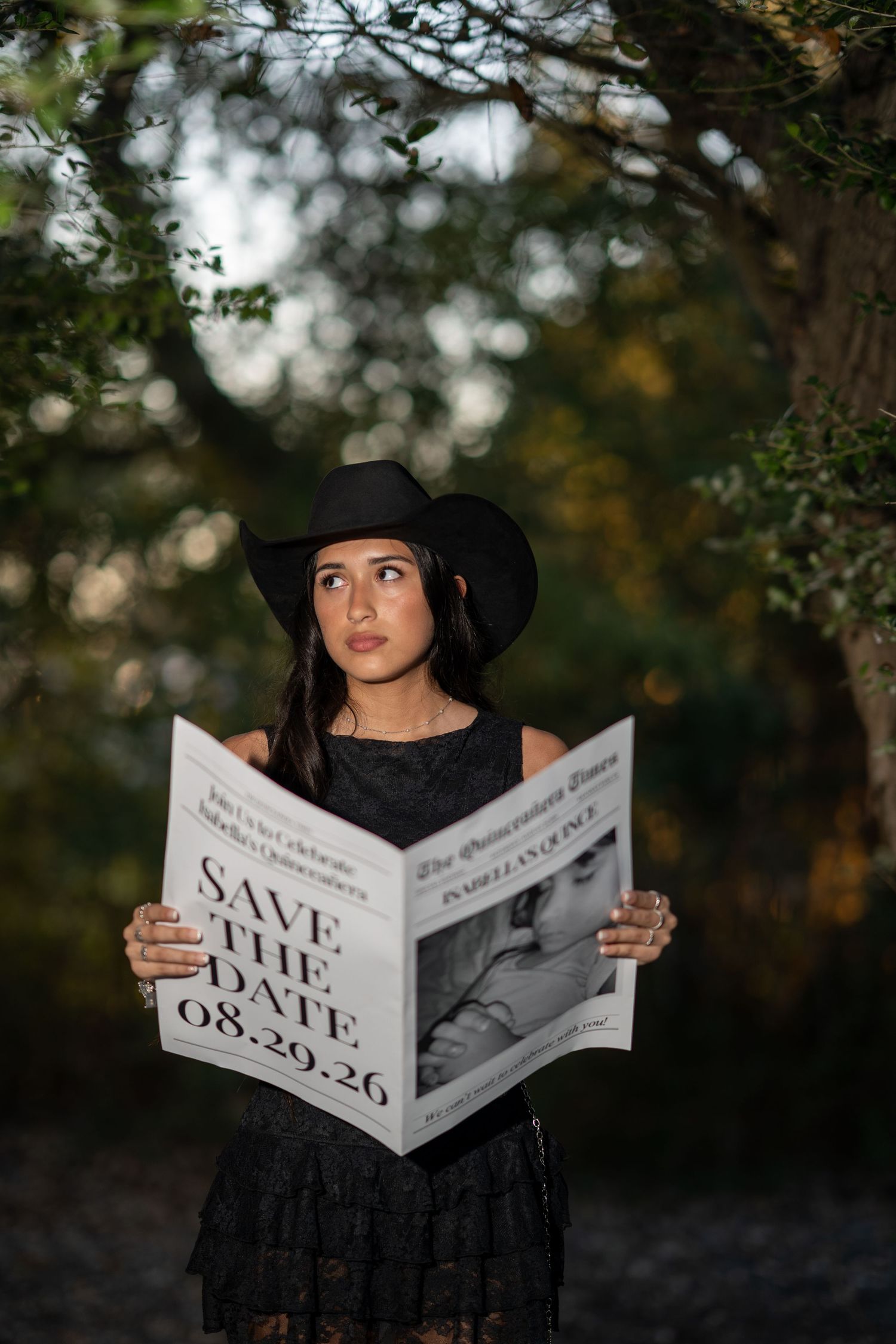 Young woman in black cowboy hat holds Save the Date newspaper announcement for 08-29-26 quinceañera outdoors.