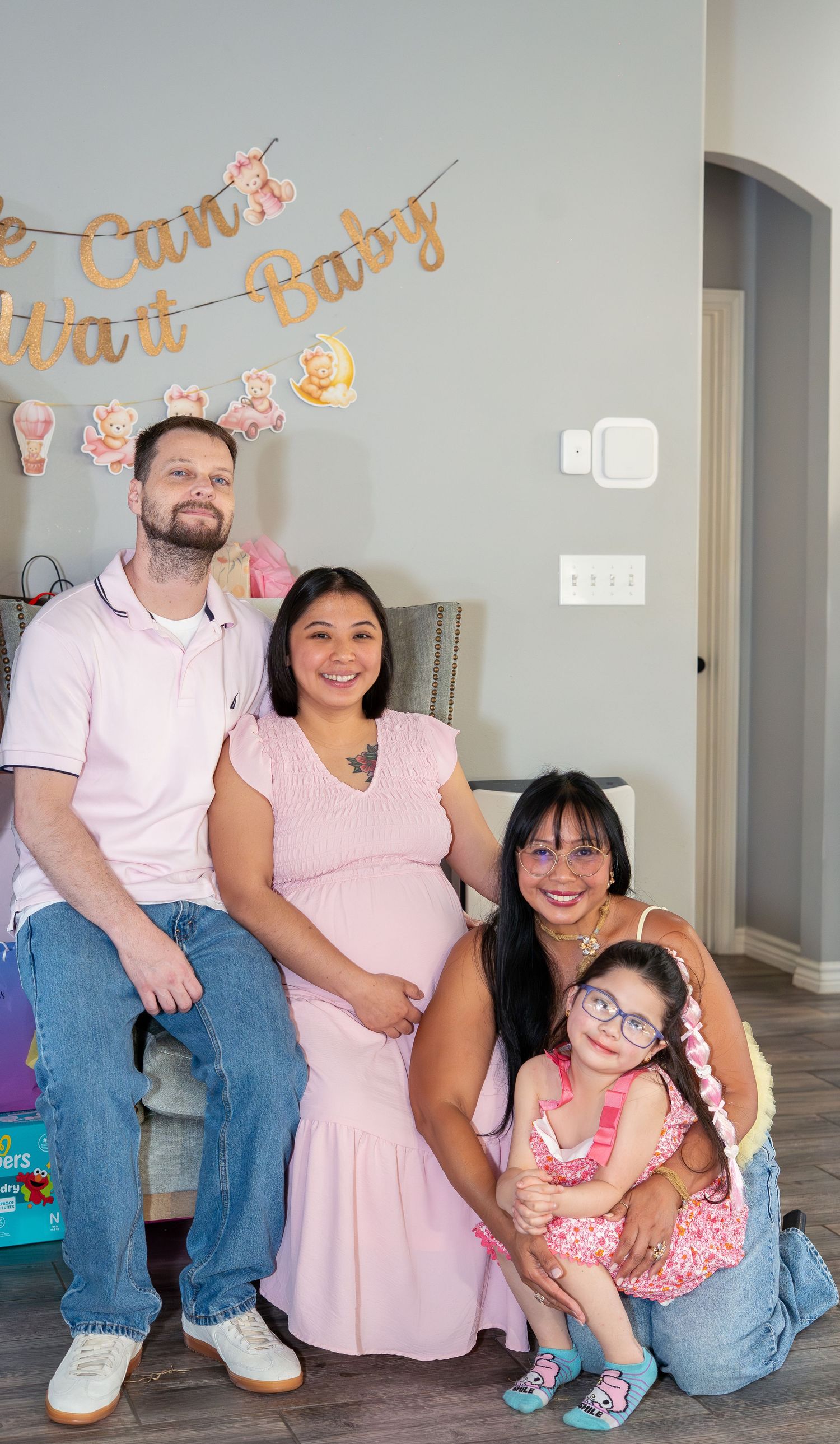 Family posing at a baby shower with a We Can't Wait Baby banner, featuring a pregnant woman in a pink dress.