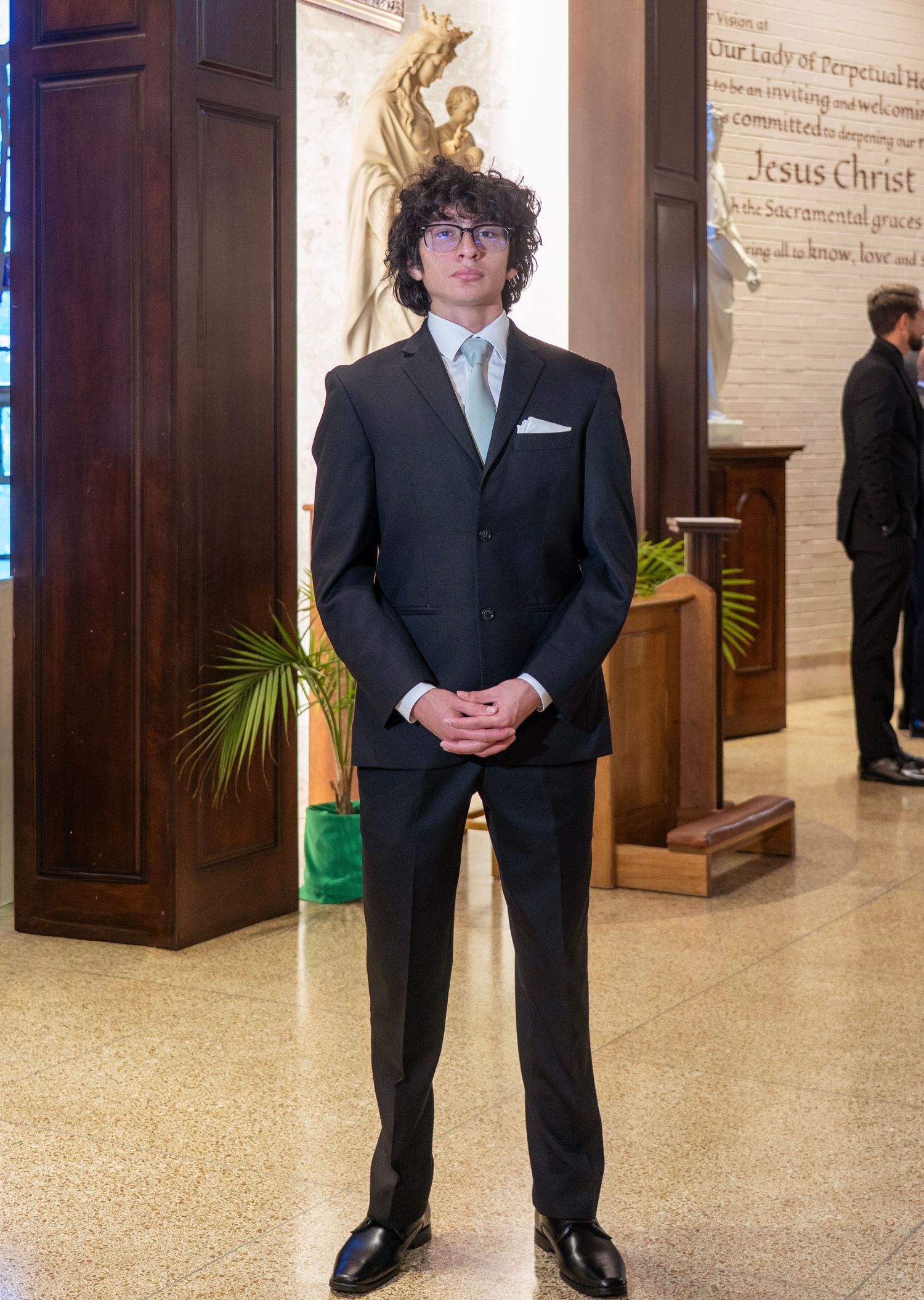 A person in a black suit stands in an elegant hallway with wooden doors and decorative wall panels.