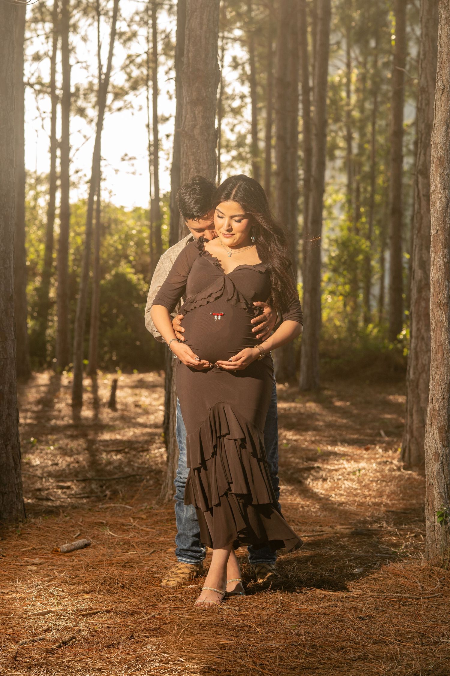 Couple sharing a tender maternity moment in a sunlit pine forest, woman in brown ruffled dress cradling baby bump.
