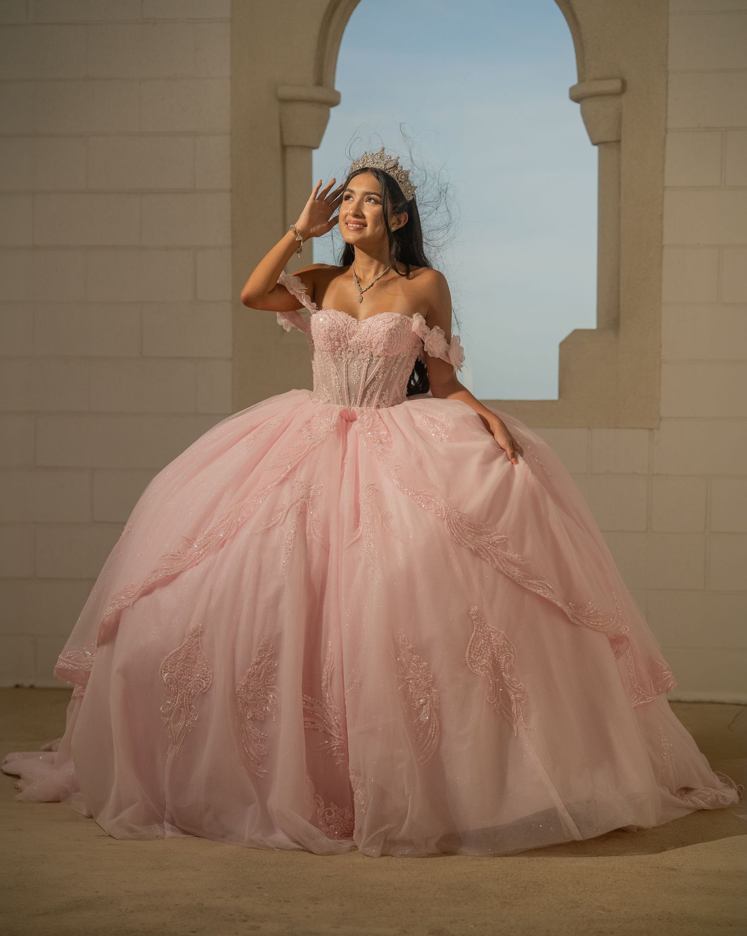 Young woman in pink off-shoulder ball gown and ornate crown poses elegantly in arched doorway.