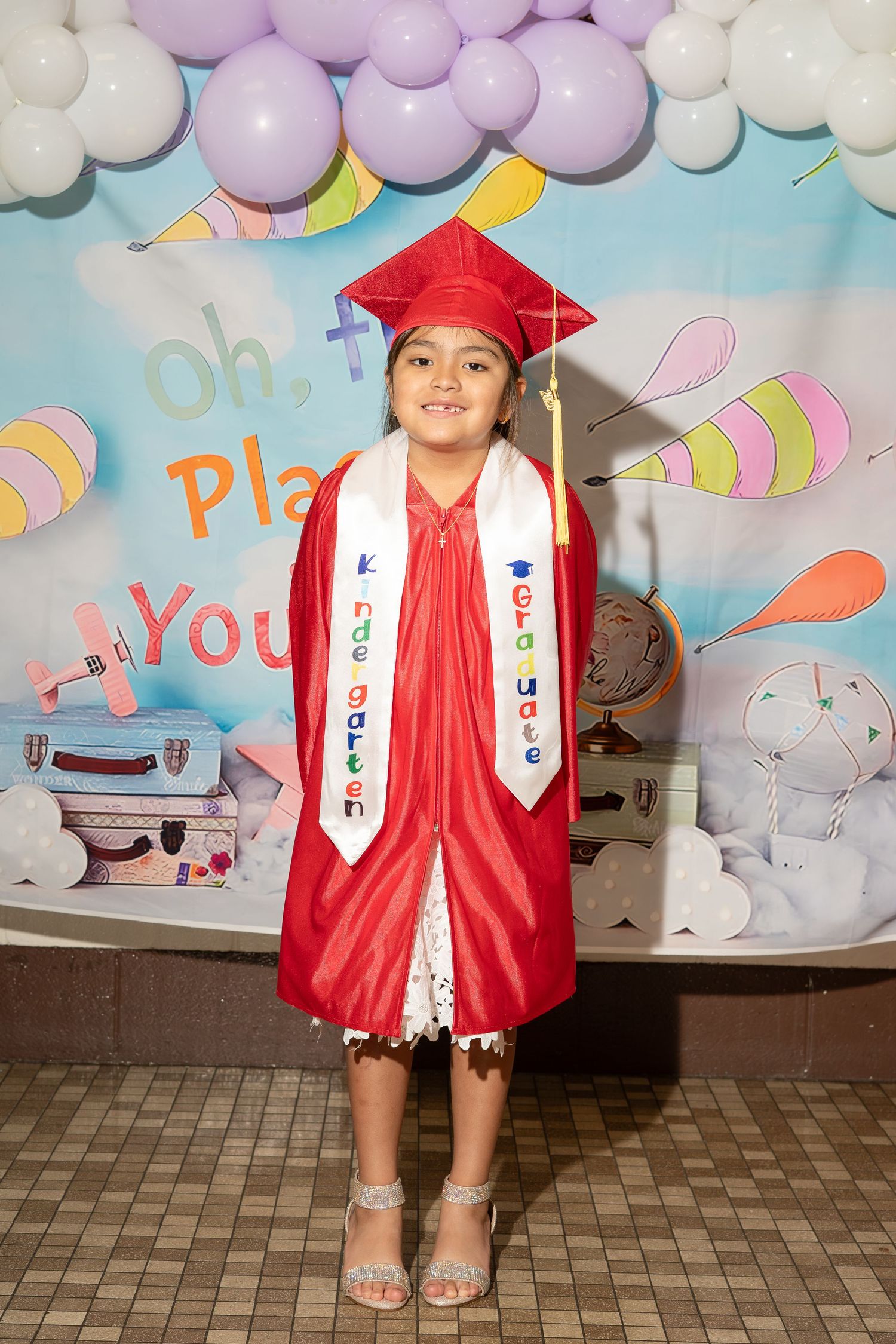 Young girl in red graduation cap and gown at kindergarten graduation ceremony with balloon backdrop.