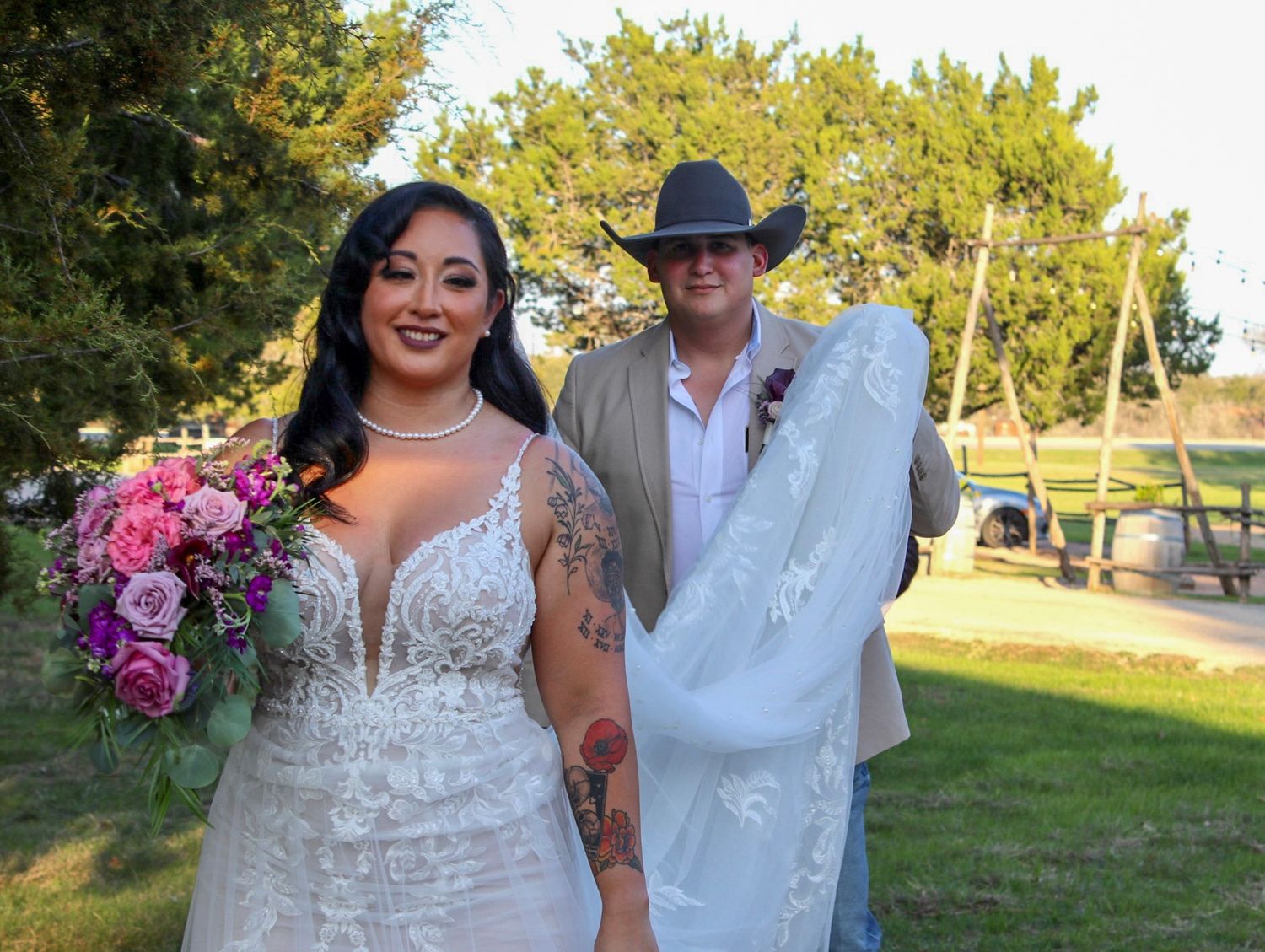 A bride in a white lace dress holds a pink bouquet while standing with someone wearing a white suit and cowboy hat.