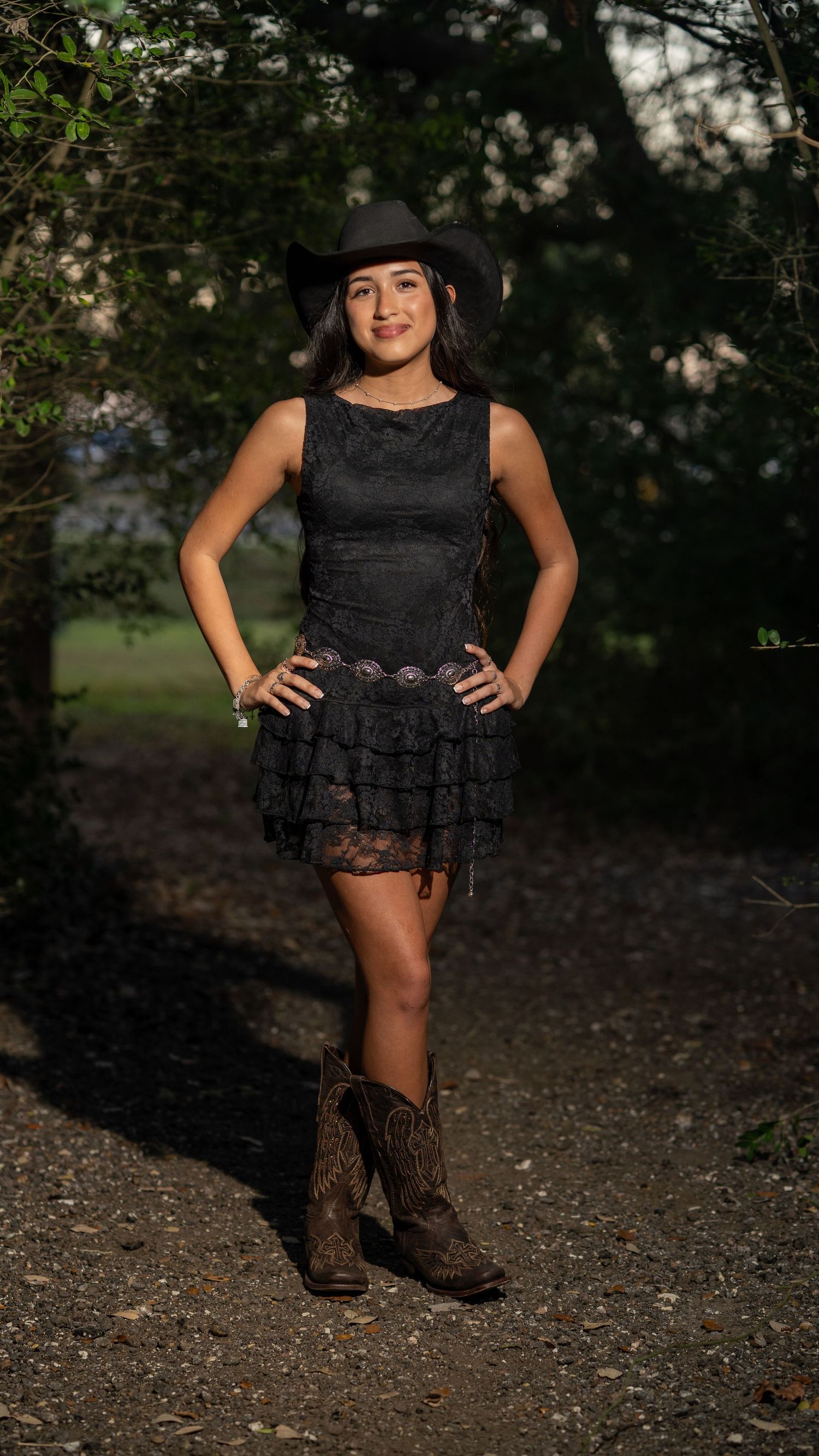 Young woman in black lace dress and cowboy boots posing confidently on a shaded outdoor path.