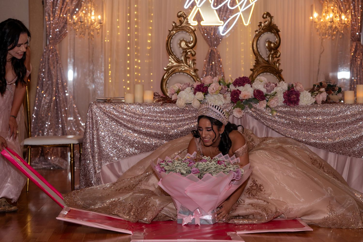 Young woman in a gold quinceañera ball gown wearing a tiara sits inside a giant pink flower prop at a celebration.