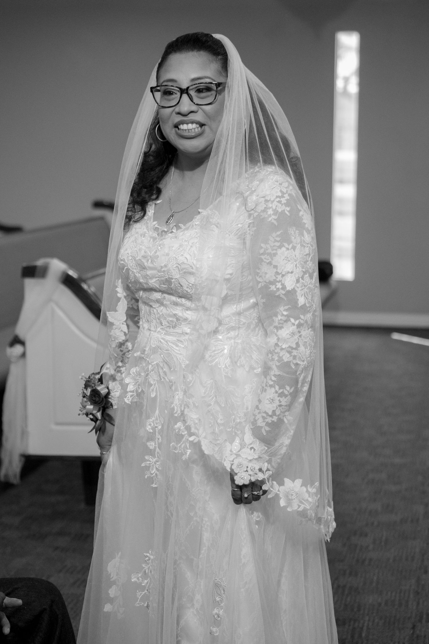 Bride in lace long-sleeve wedding dress and veil smiles in black and white church portrait.