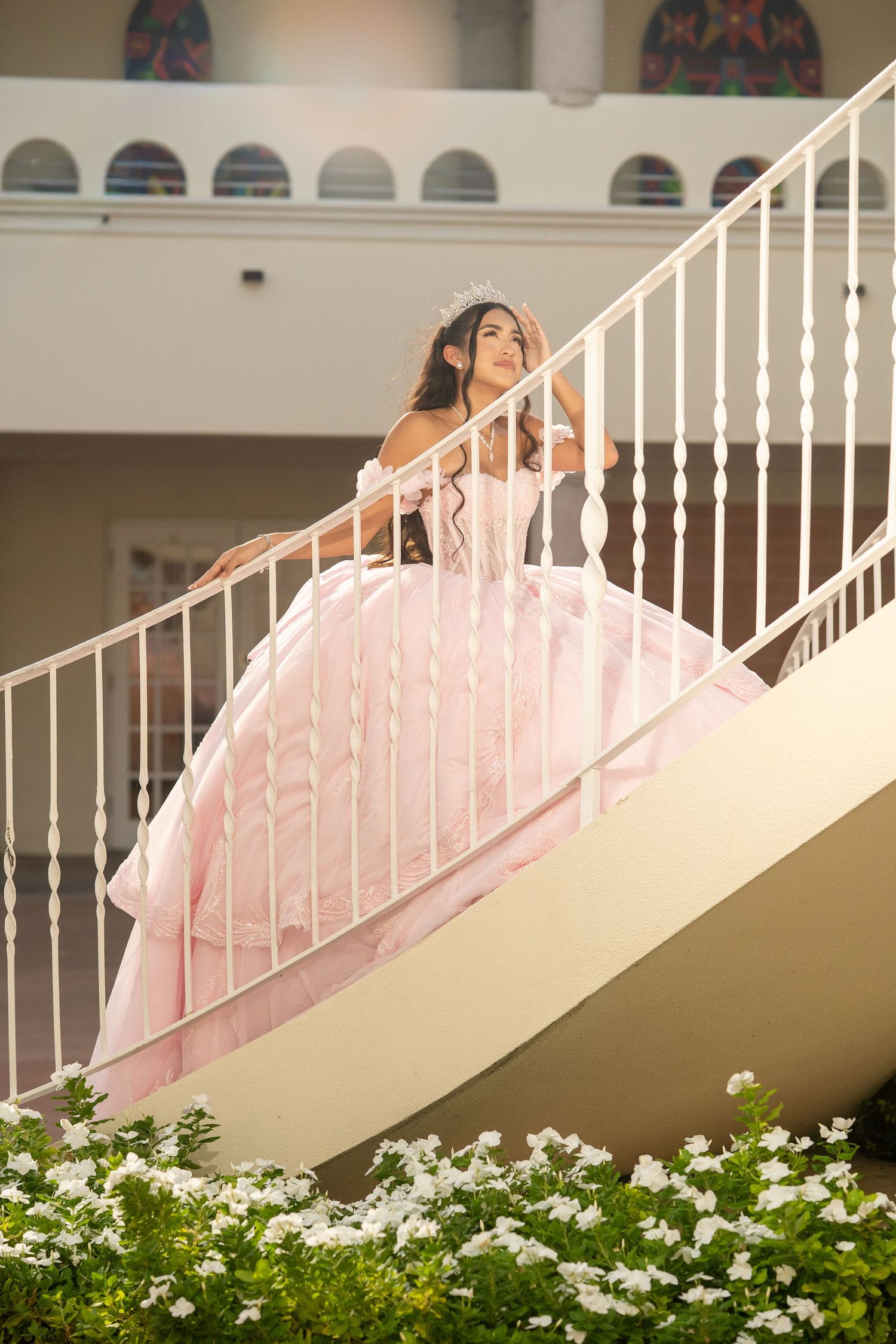 Young woman in a pink ball gown and tiara poses gracefully on white staircase during quinceañera celebration.