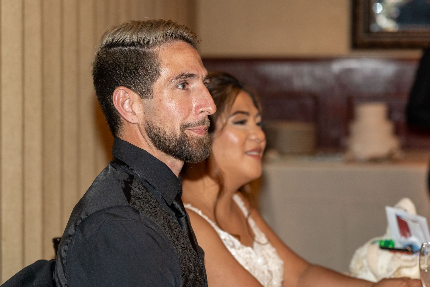 A man in a black shirt and woman in a lace wedding dress sit together during a wedding ceremony.