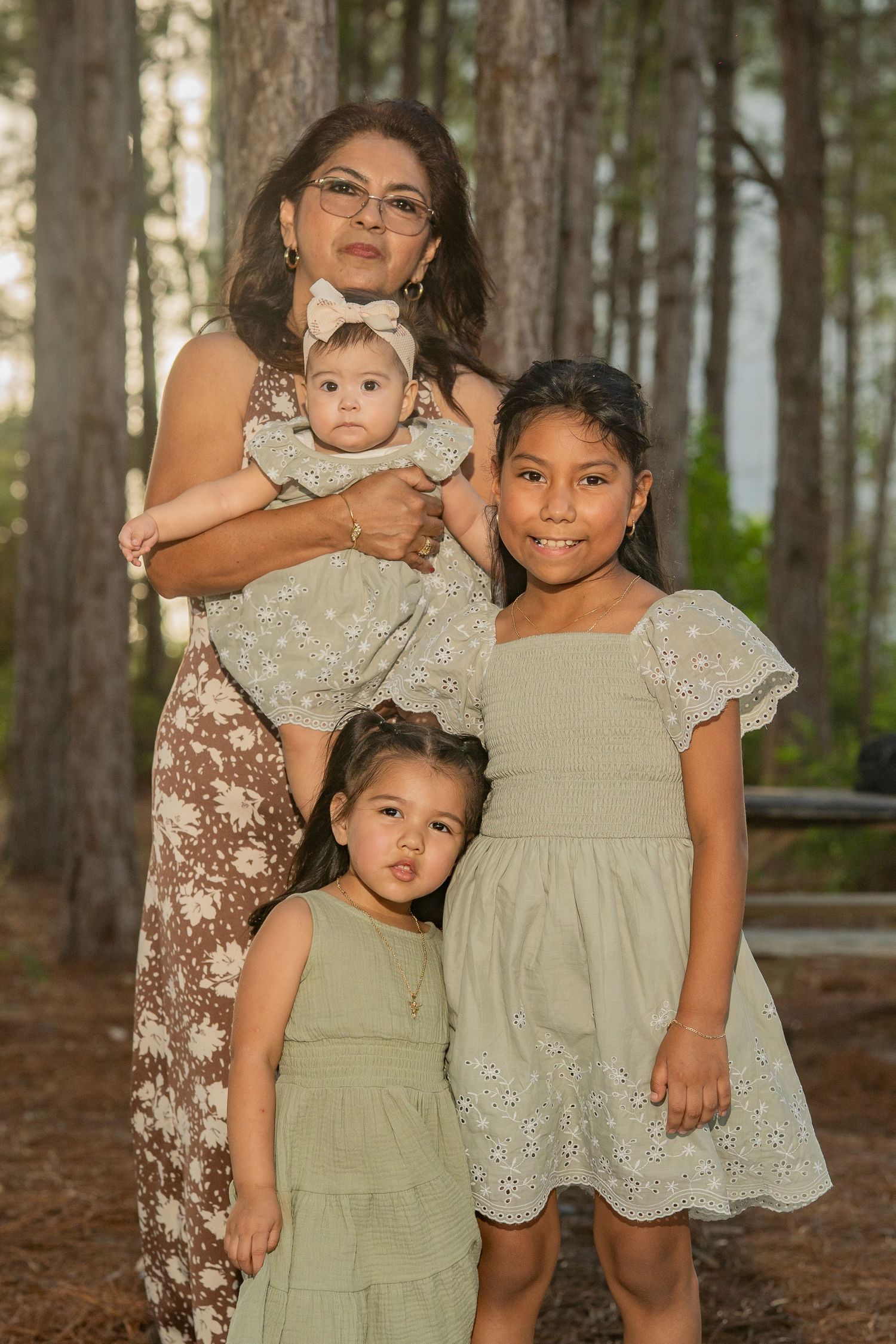 A grandmother holds a baby while posing with two young girls in matching sage green dresses among pine trees.
