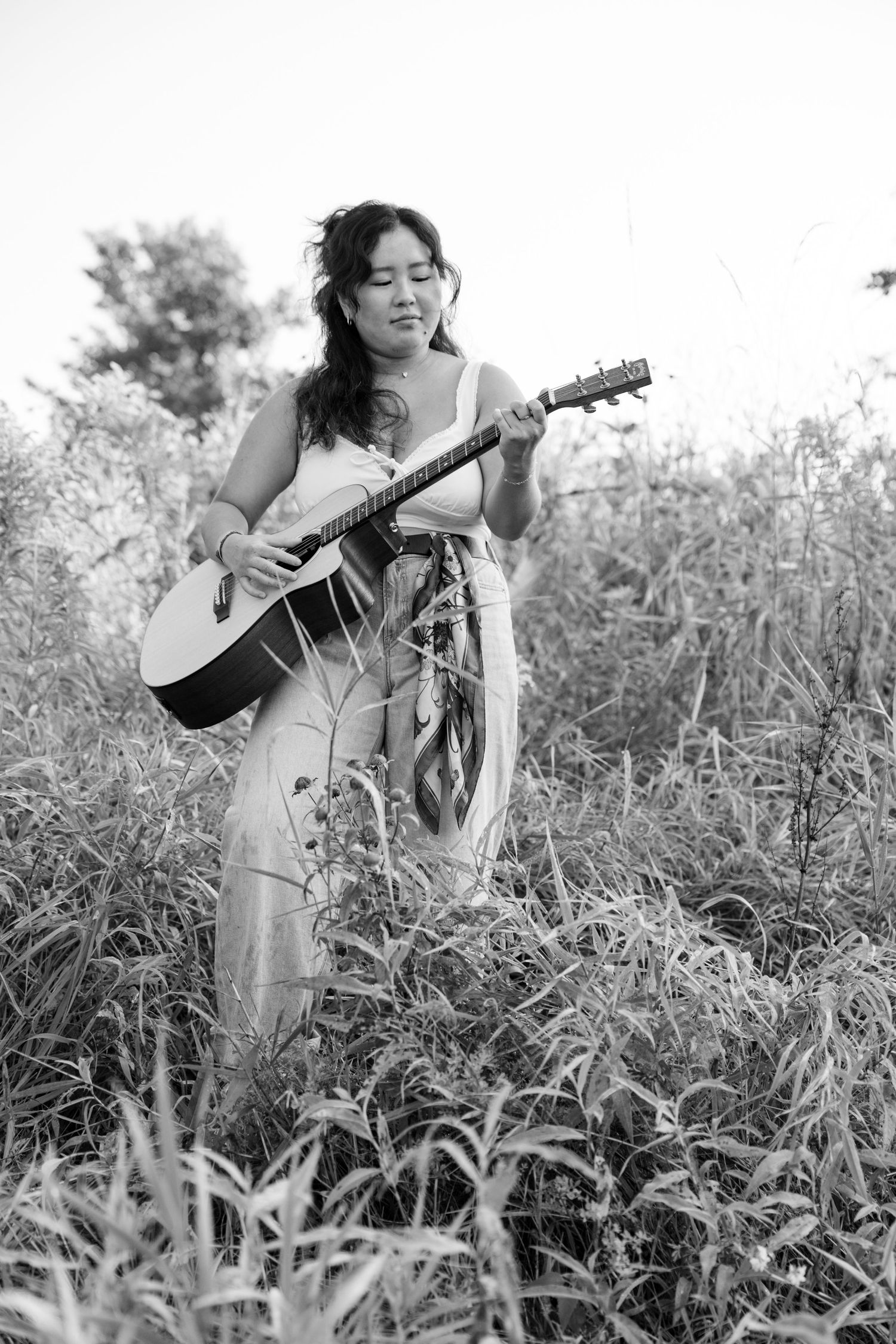 Person playing acoustic guitar while standing in tall grass field in black and white photograph.