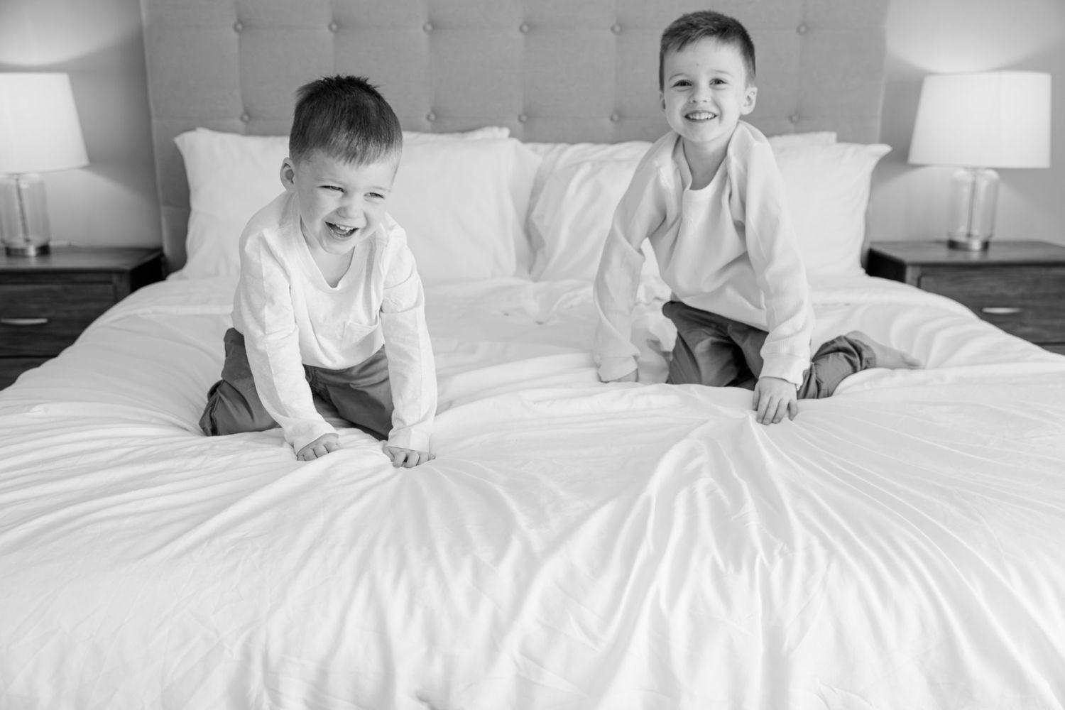 Two children laughing together on white bed in bright bedroom setting.