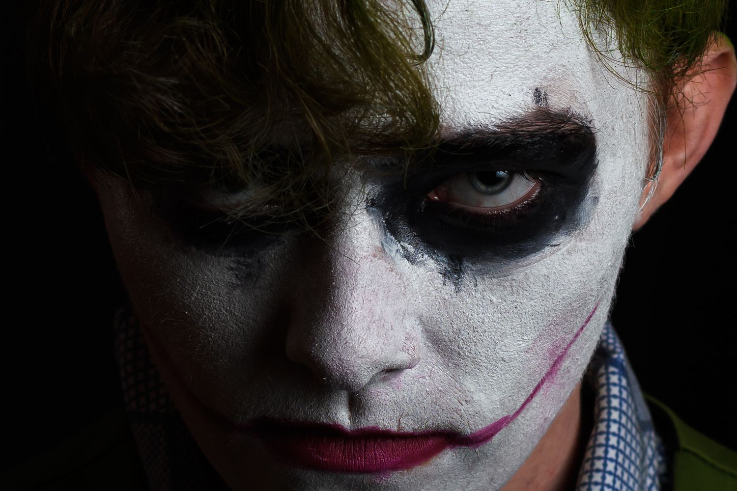 Close-up of someone in dramatic Joker-style Halloween makeup with dark eye paint and red lipstick against dark background.