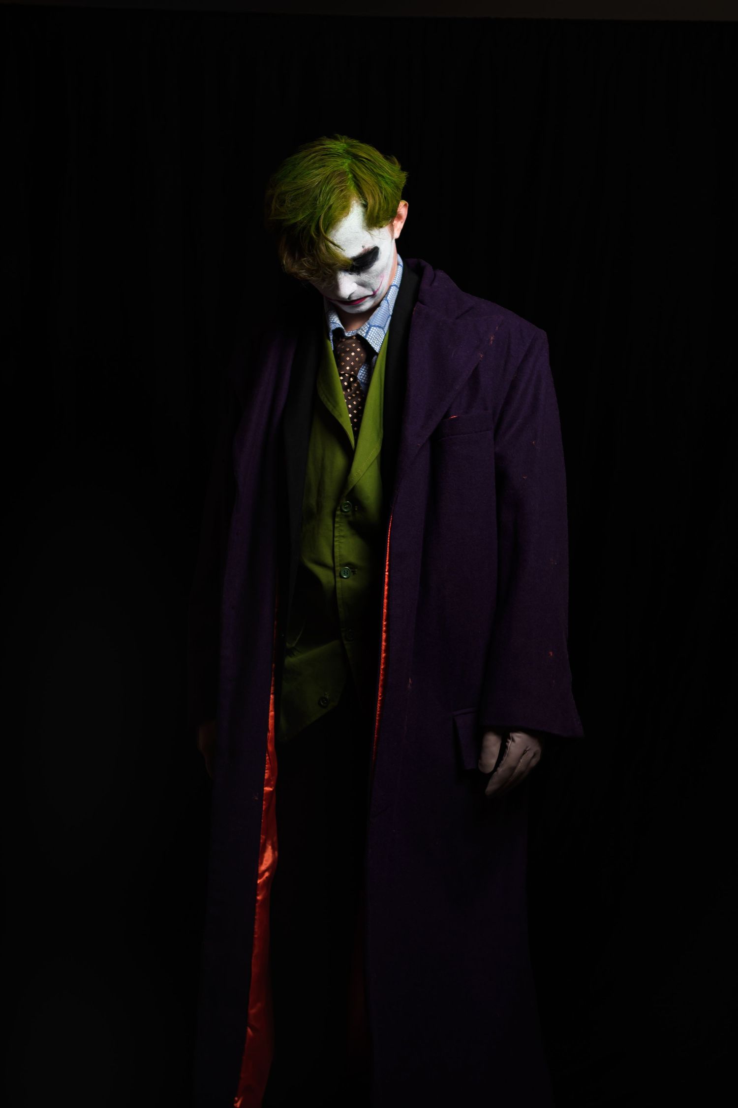A dark and moody cosplay featuring dramatic purple coat and green clothing against a black background.