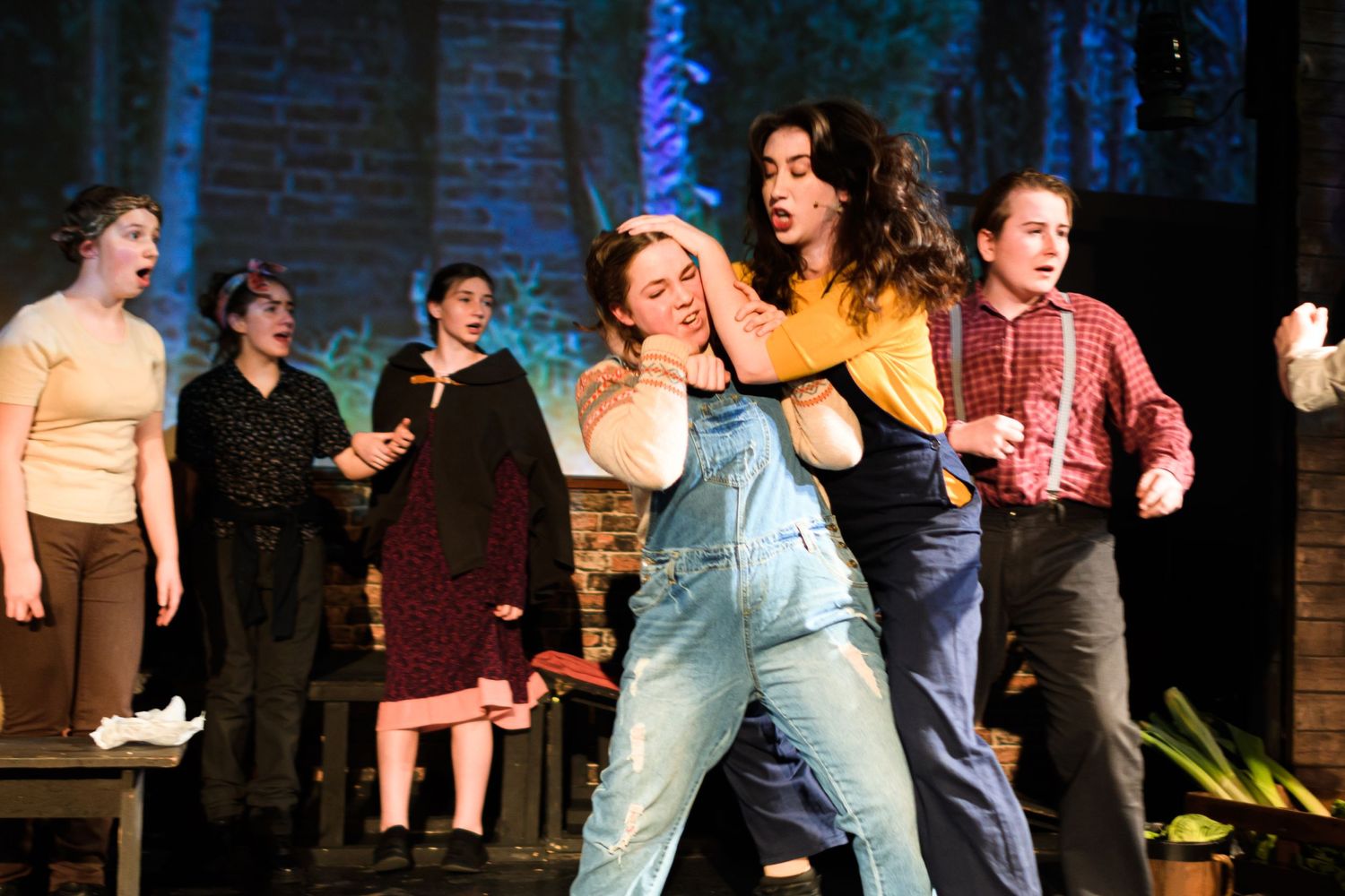 Energetic theatrical performance with ensemble cast dancing in front of brick wall set.