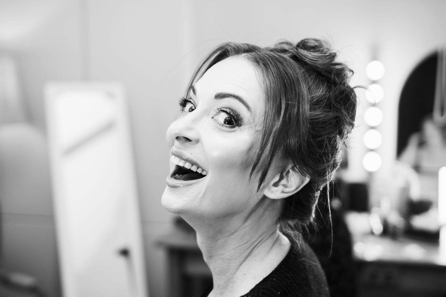A woman with dramatic makeup laughs joyfully while looking upward in a black and white portrait.