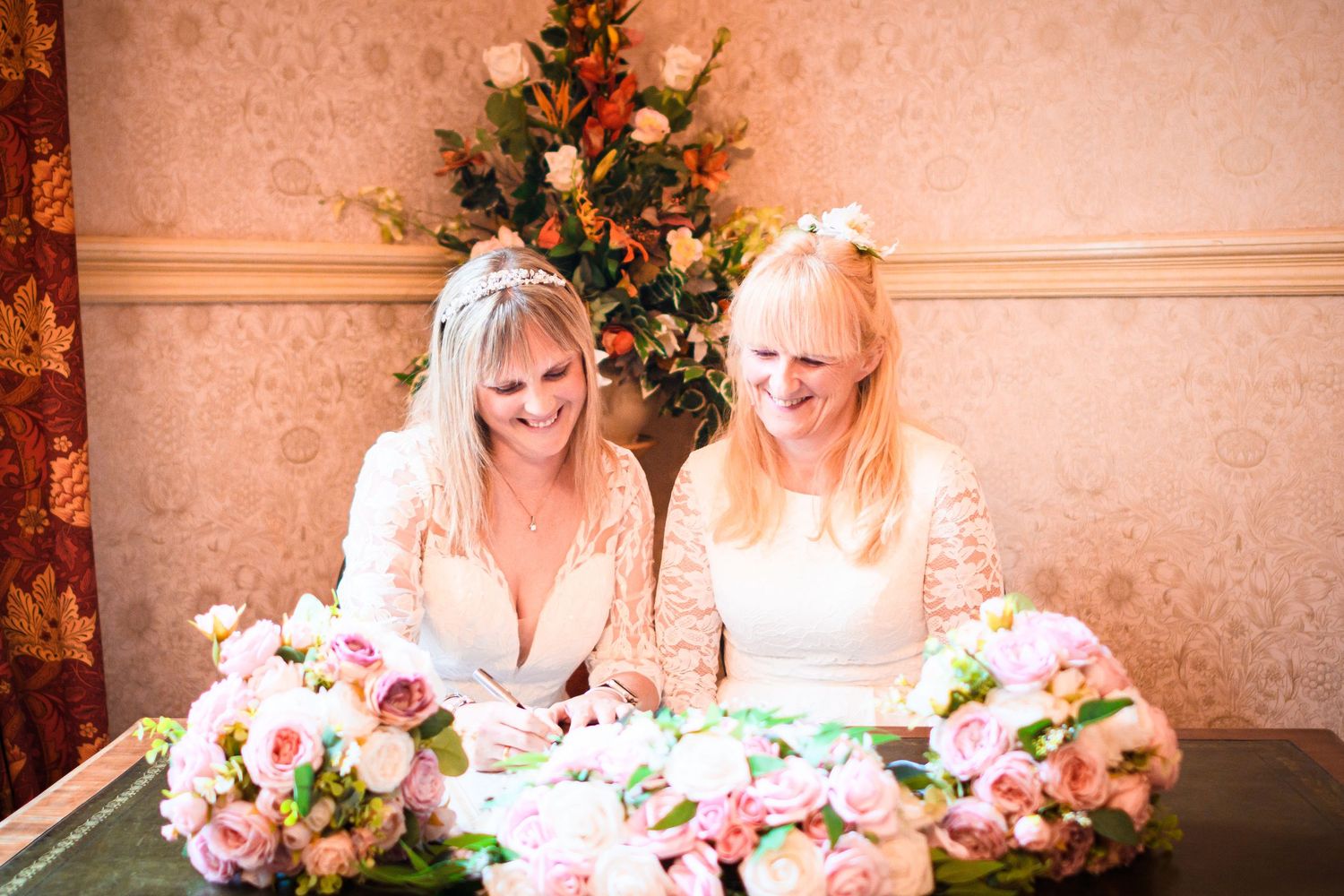 Two people in white lace dresses sit together by a Christmas tree with pink floral bouquets.