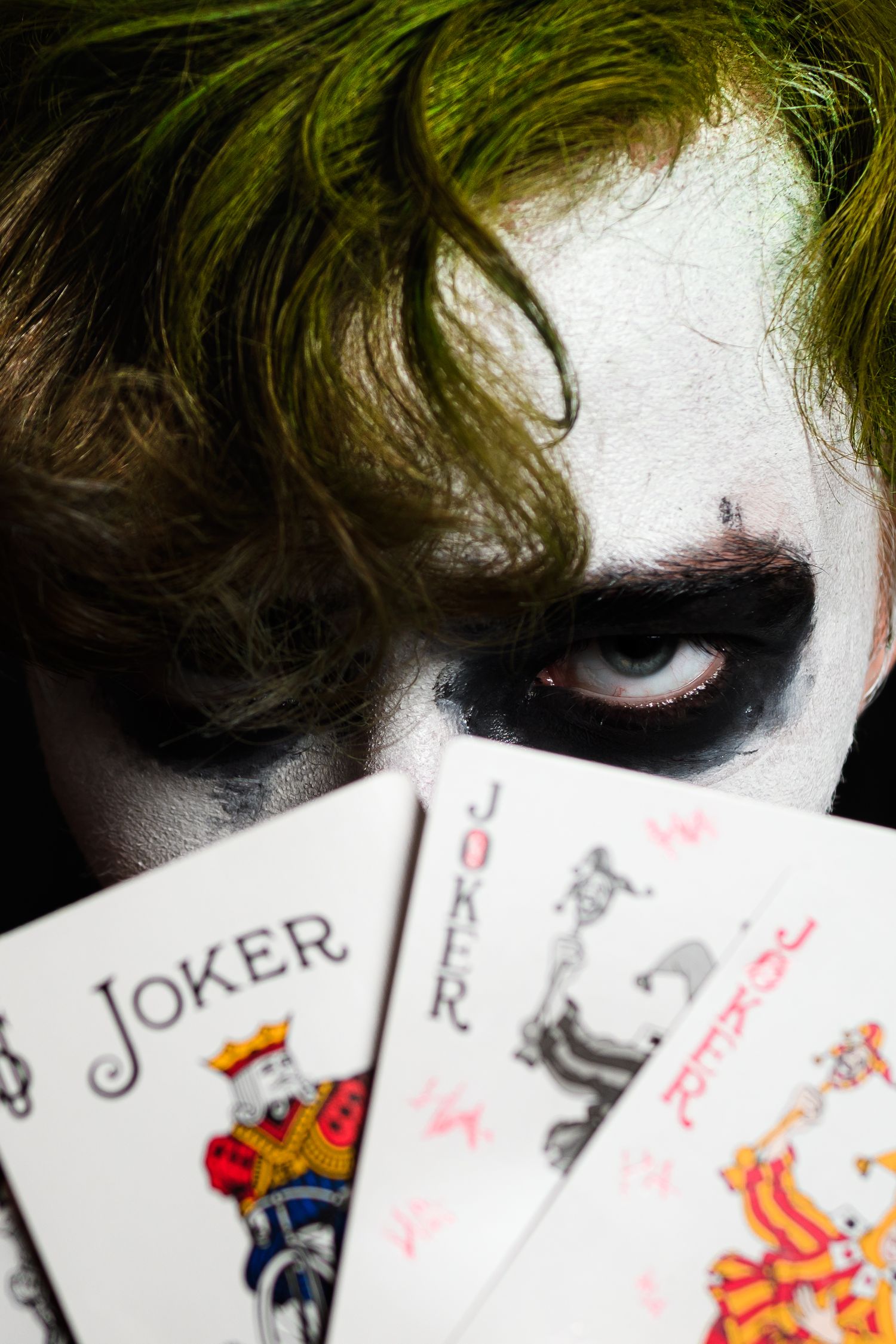 Dark clown makeup and green hair visible above playing cards showing Joker card in dramatic closeup shot.