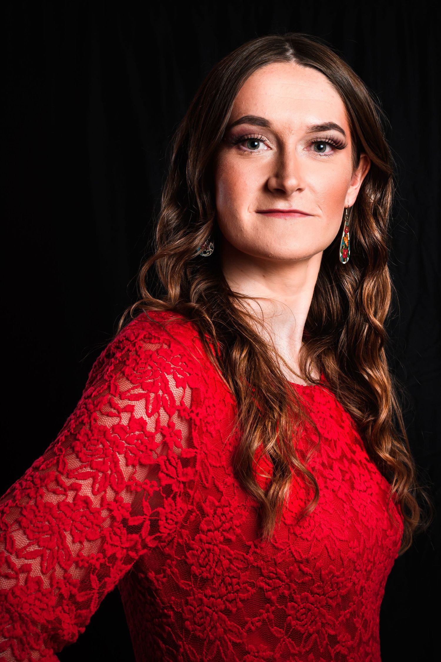 Elegant portrait in red lace dress against dark background with long wavy hair.