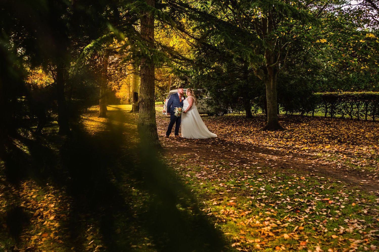 A romantic autumn wedding photo captures newlyweds walking through a golden-lit wooded path with fallen leaves.