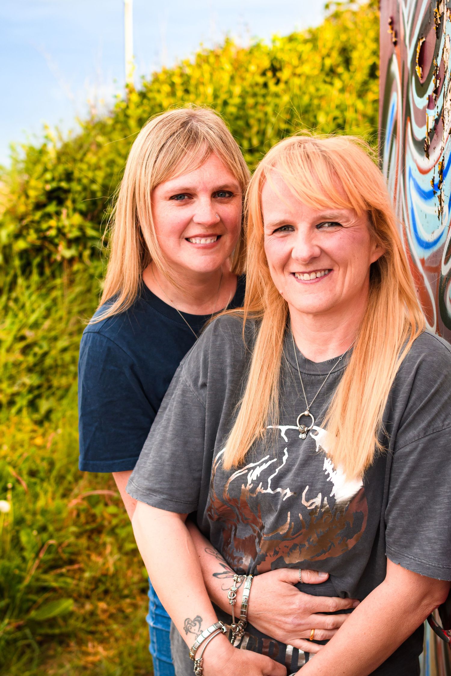 Two blonde women in casual t-shirts smile warmly outdoors against green foliage background.