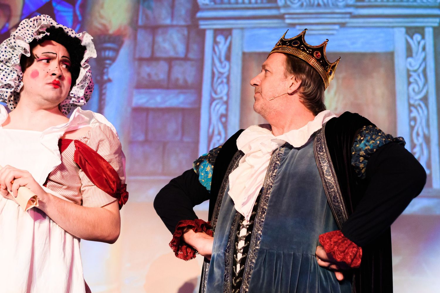 Two performers in ornate renaissance costumes act in a scene with elaborate backdrop.