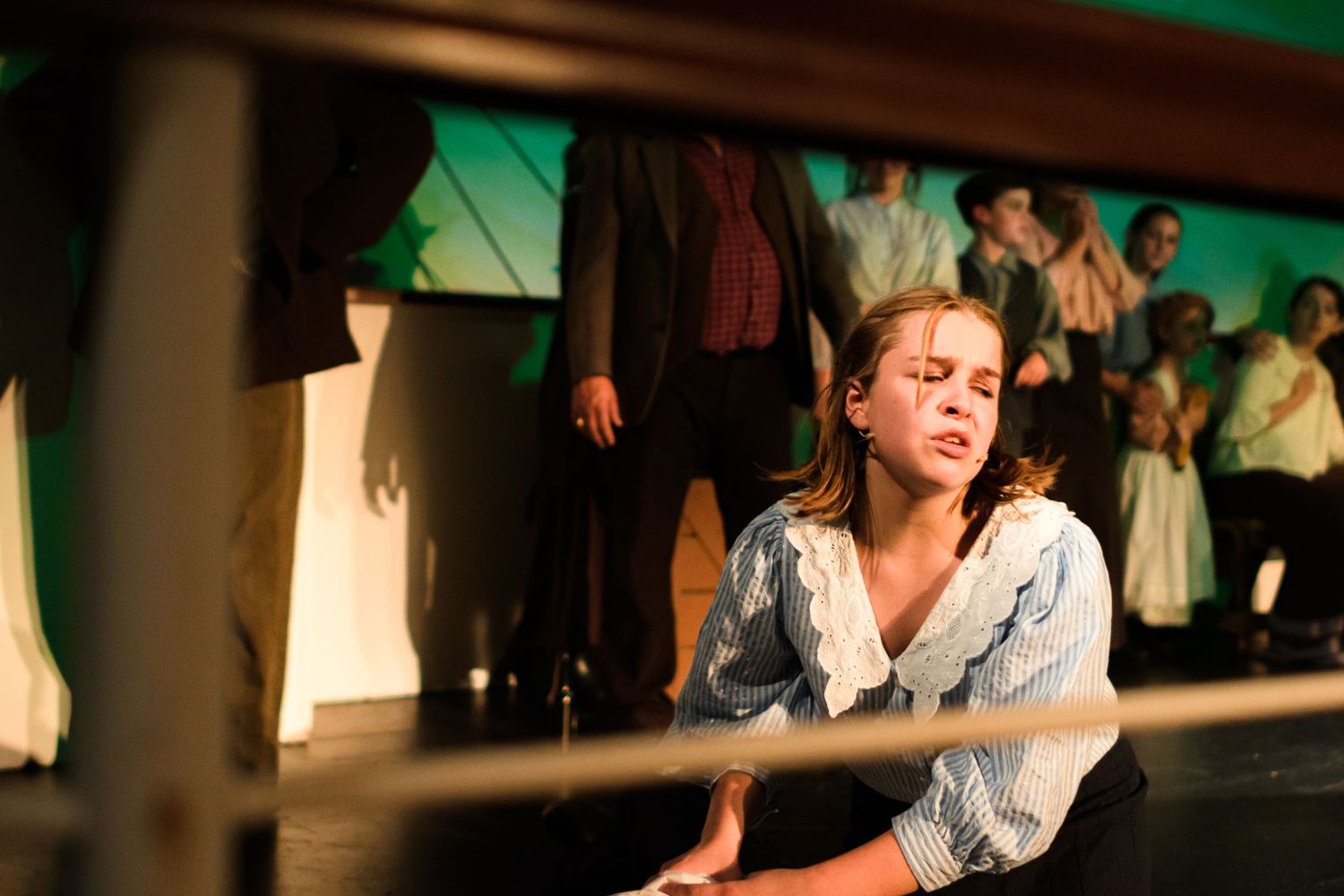 A performer in vintage clothing leans forward during a theatrical scene with other actors in the background.