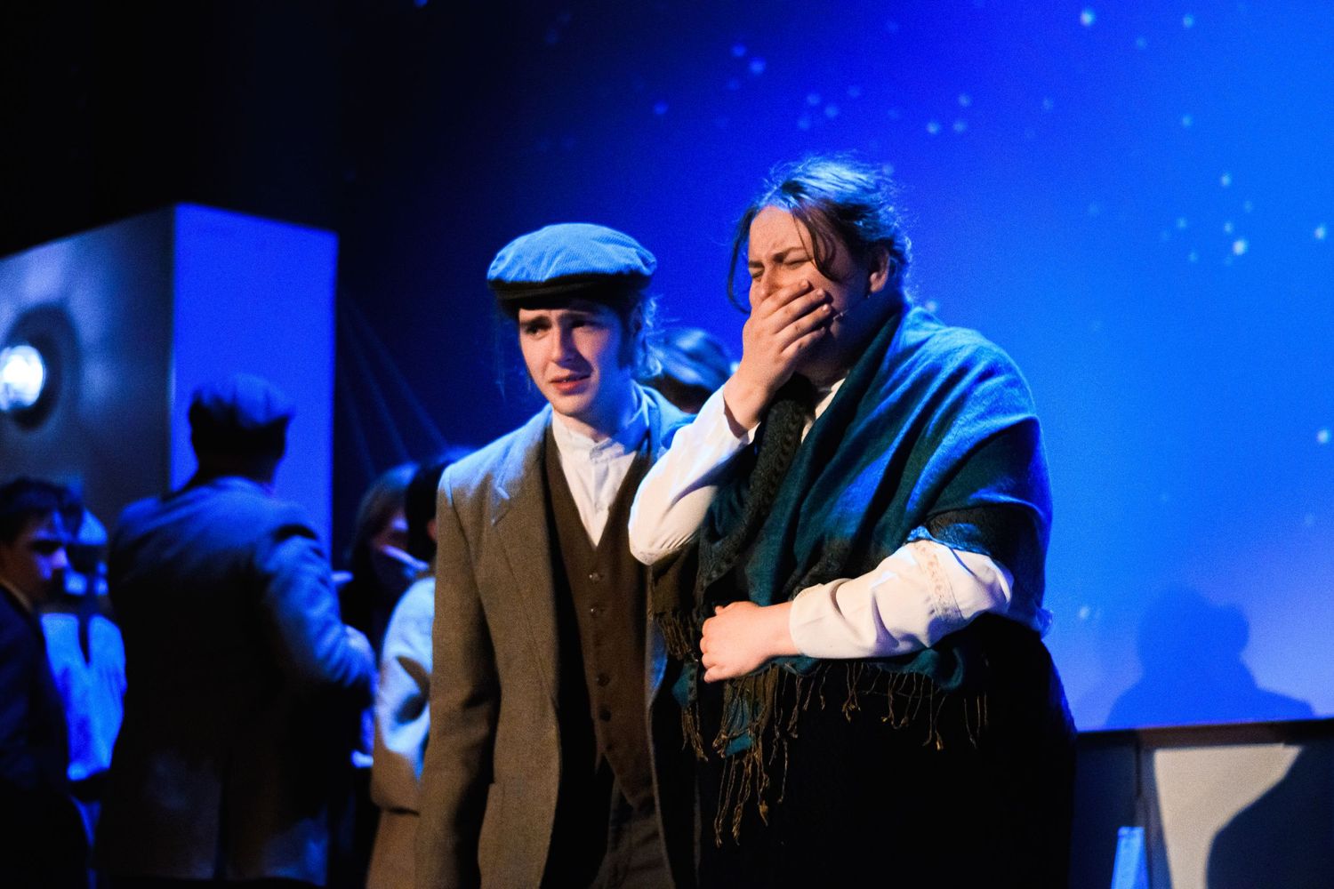 Two performers in period costumes react emotionally during a stage performance with blue lighting.