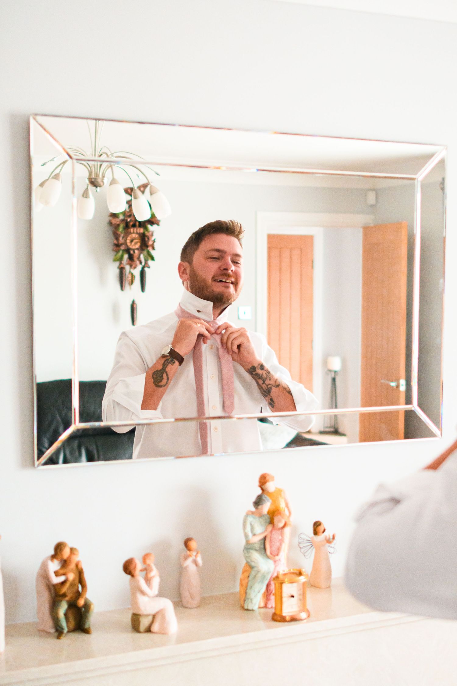 Reflection of person adjusting pink bowtie in mirror with decorative figurines on shelf.