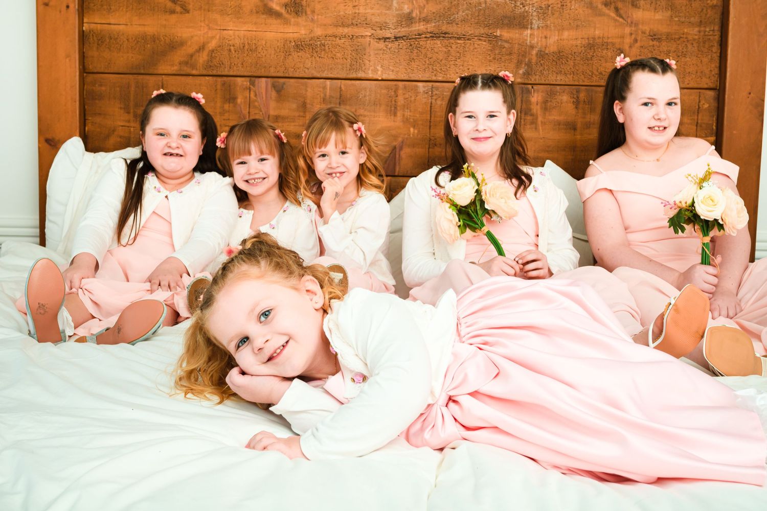 Group of young bridesmaids in pink dresses lounging together on white bedding.