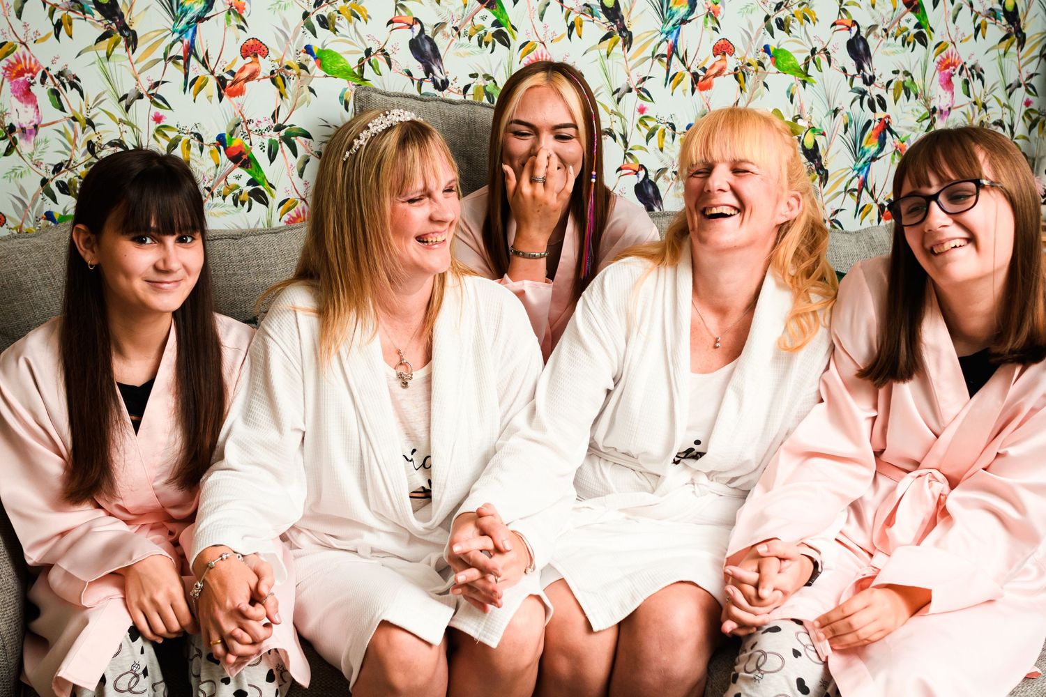 Group of bridesmaids in white and pink robes sitting together against patterned wallpaper.
