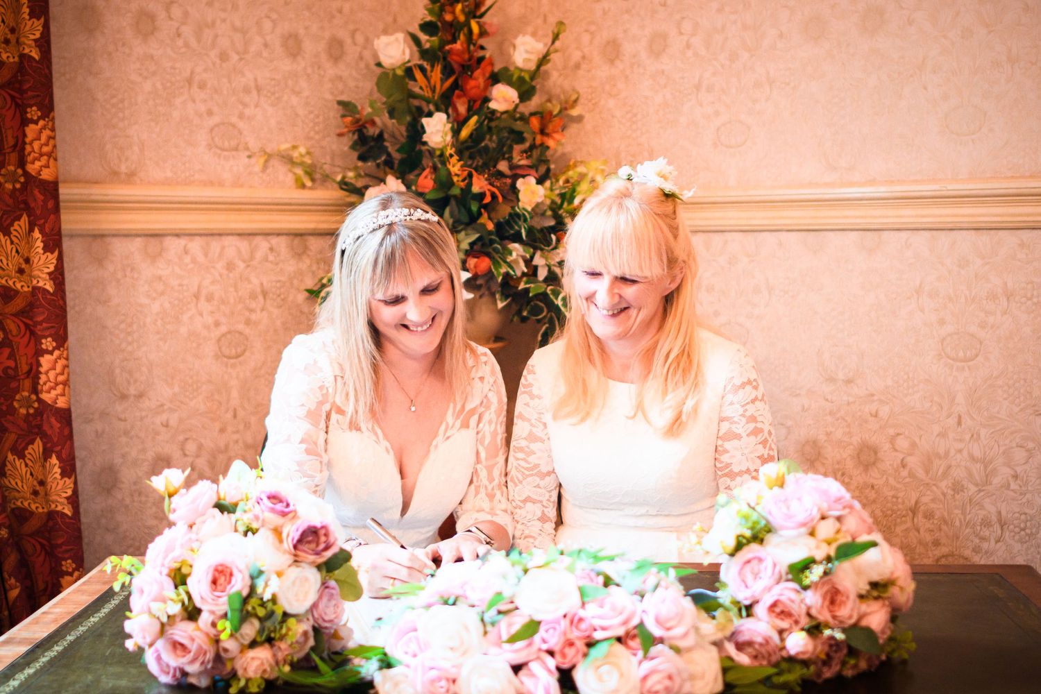 Two brides in white dresses share a moment at their reception table with decorated Christmas tree in background.