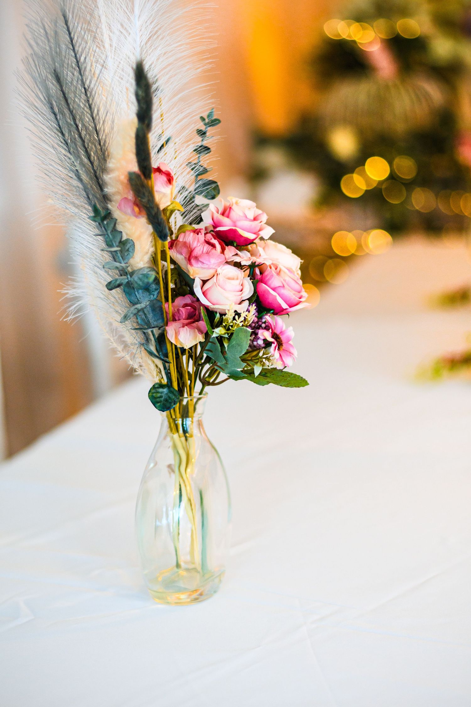 Delicate floral arrangement with pink roses and feathers in a clear glass vase against bokeh lights.