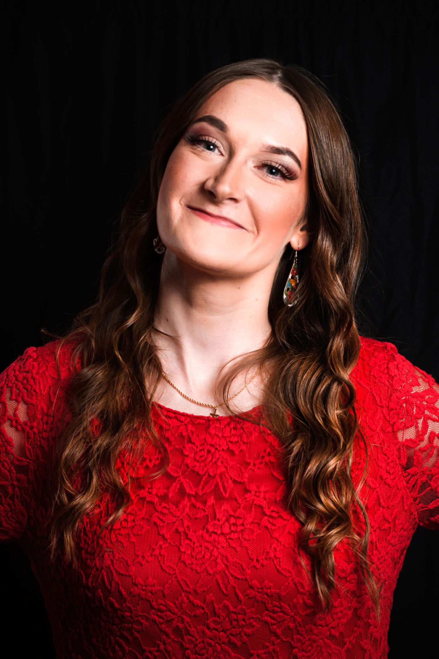 Portrait of someone in a red lace dress with long wavy brown hair against a dark background.