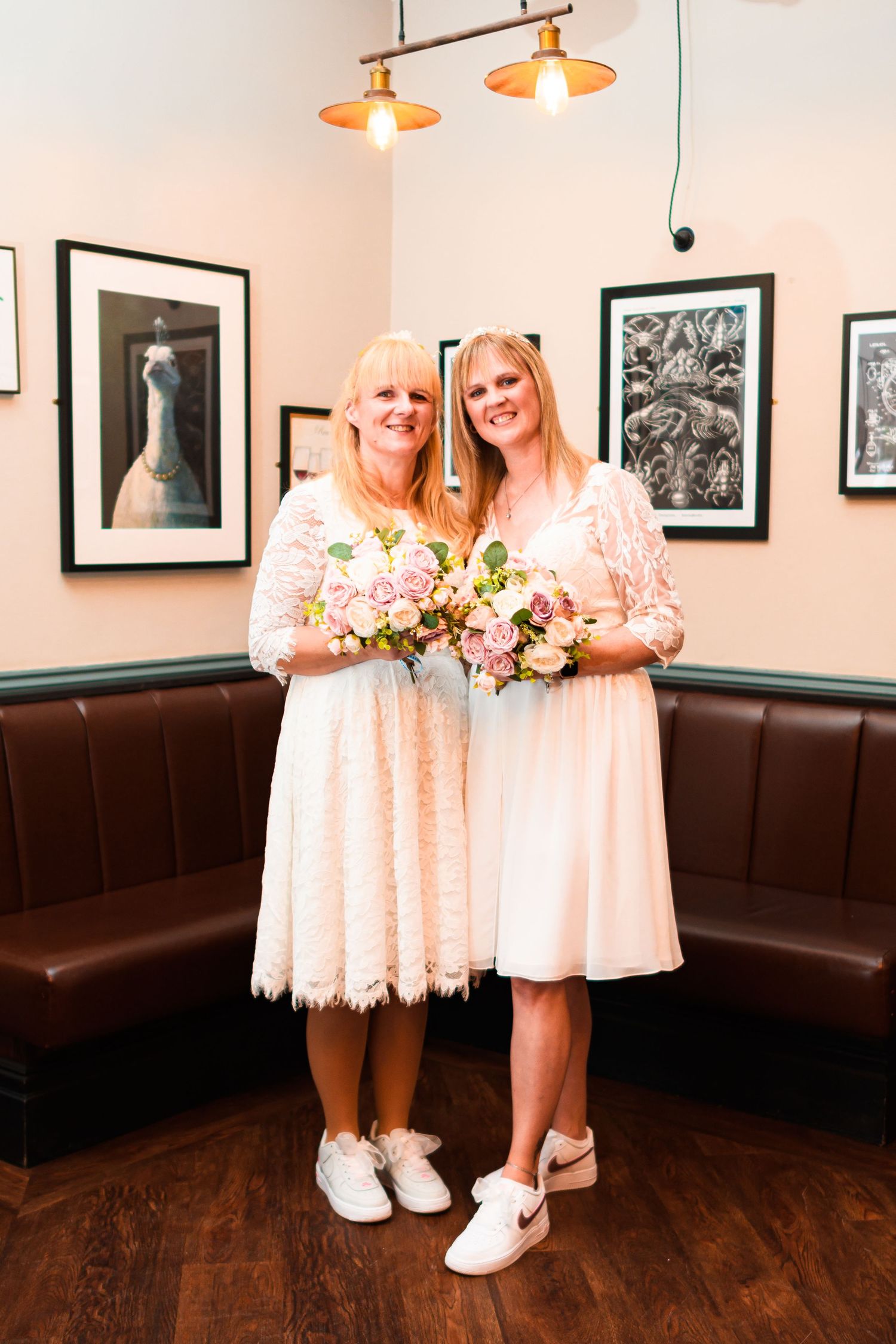 Two figures in matching peach-colored lace dresses and white shoes stand on hardwood floors near black leather booth seating.