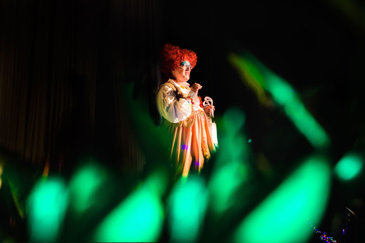A performer in traditional costume stands dramatically lit by green spotlights on stage.