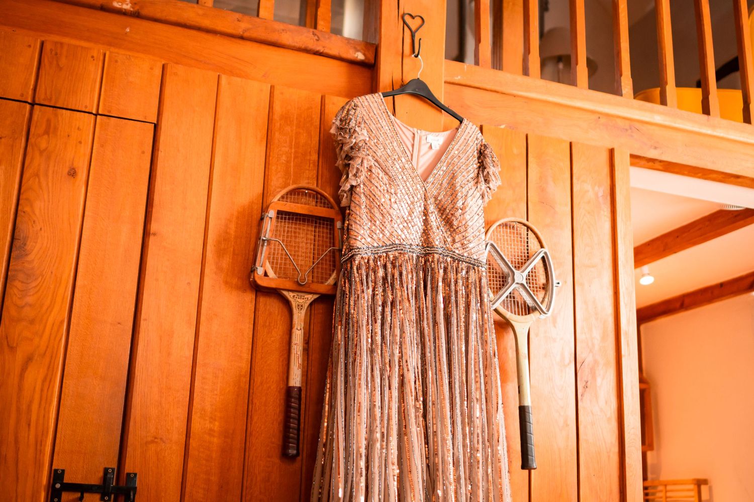 A vintage-style blush pink lace dress hangs on a wooden hanger against orange wooden paneled walls.